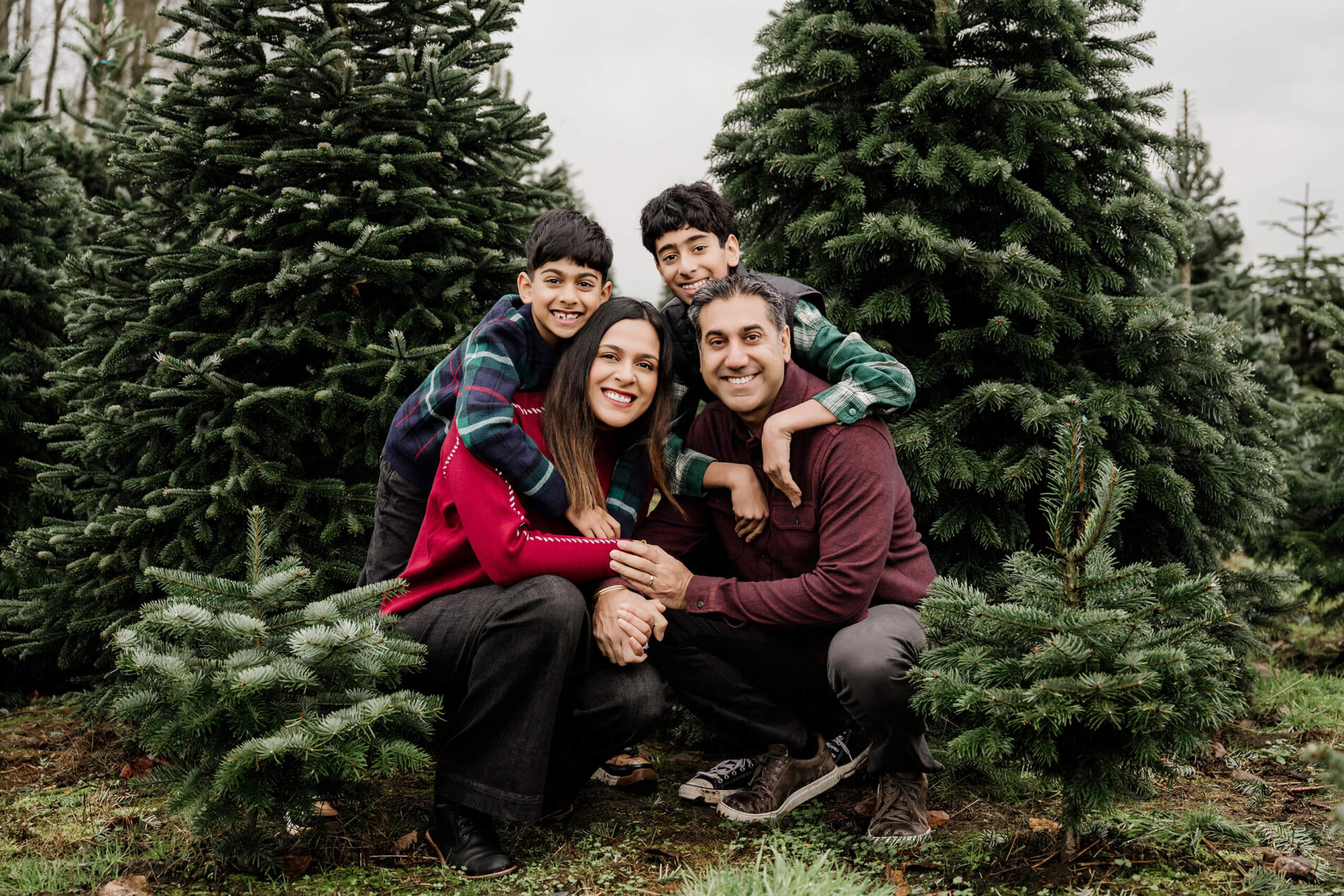 Mom and dad crouch between evergreen trees as their two sons lean over their shoulders for a family portrait.