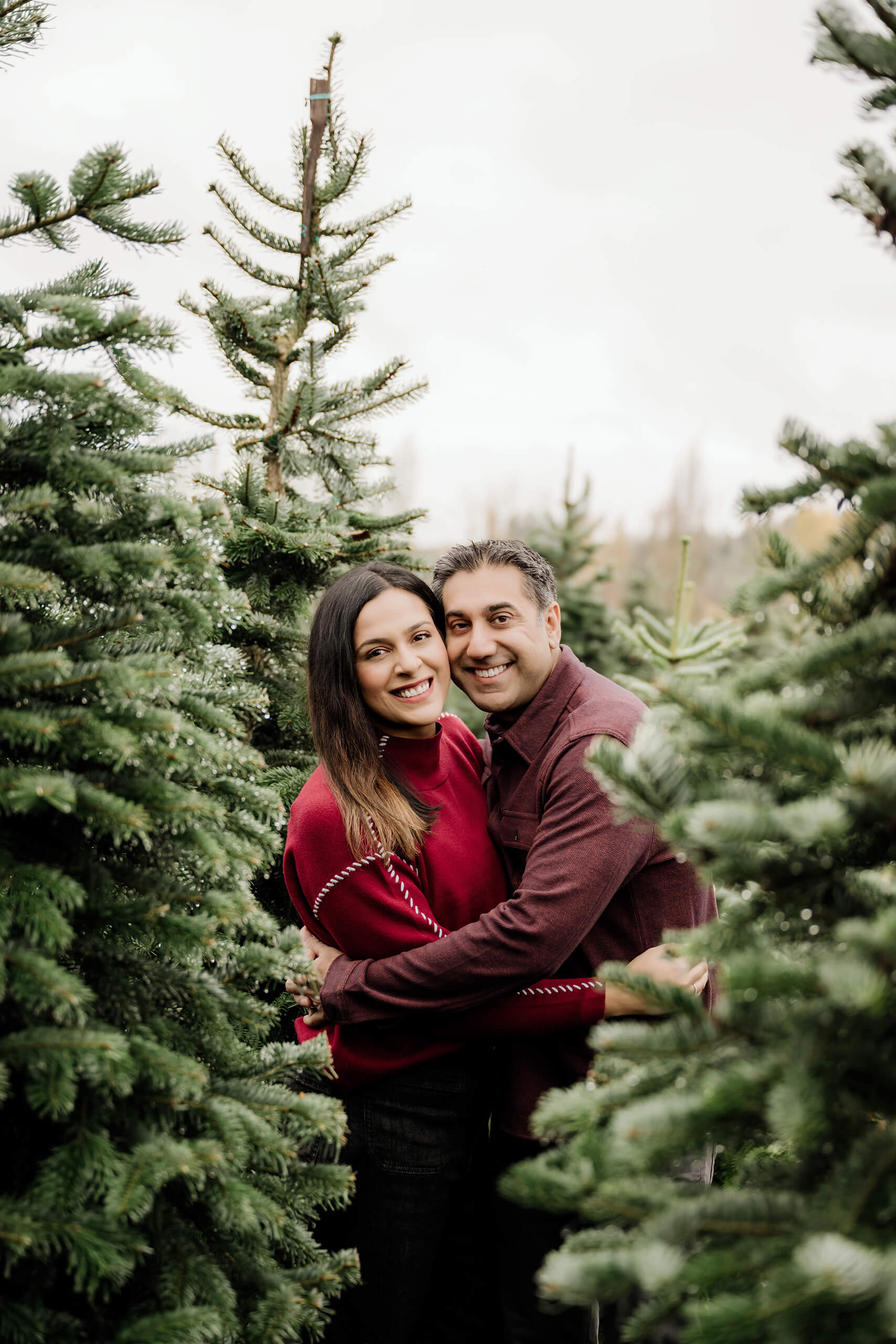 Mom and dad embrace and smile at the camera between evergreen trees.