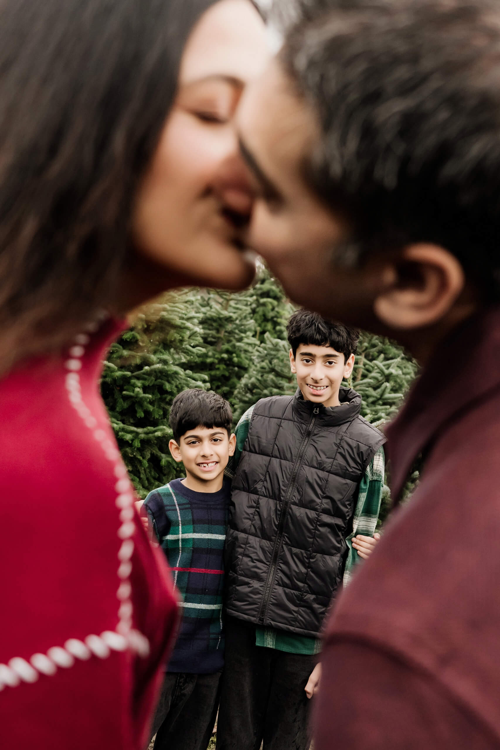Mom and dad kiss in the foreground while their two sons smile between the evergreen trees behind them.