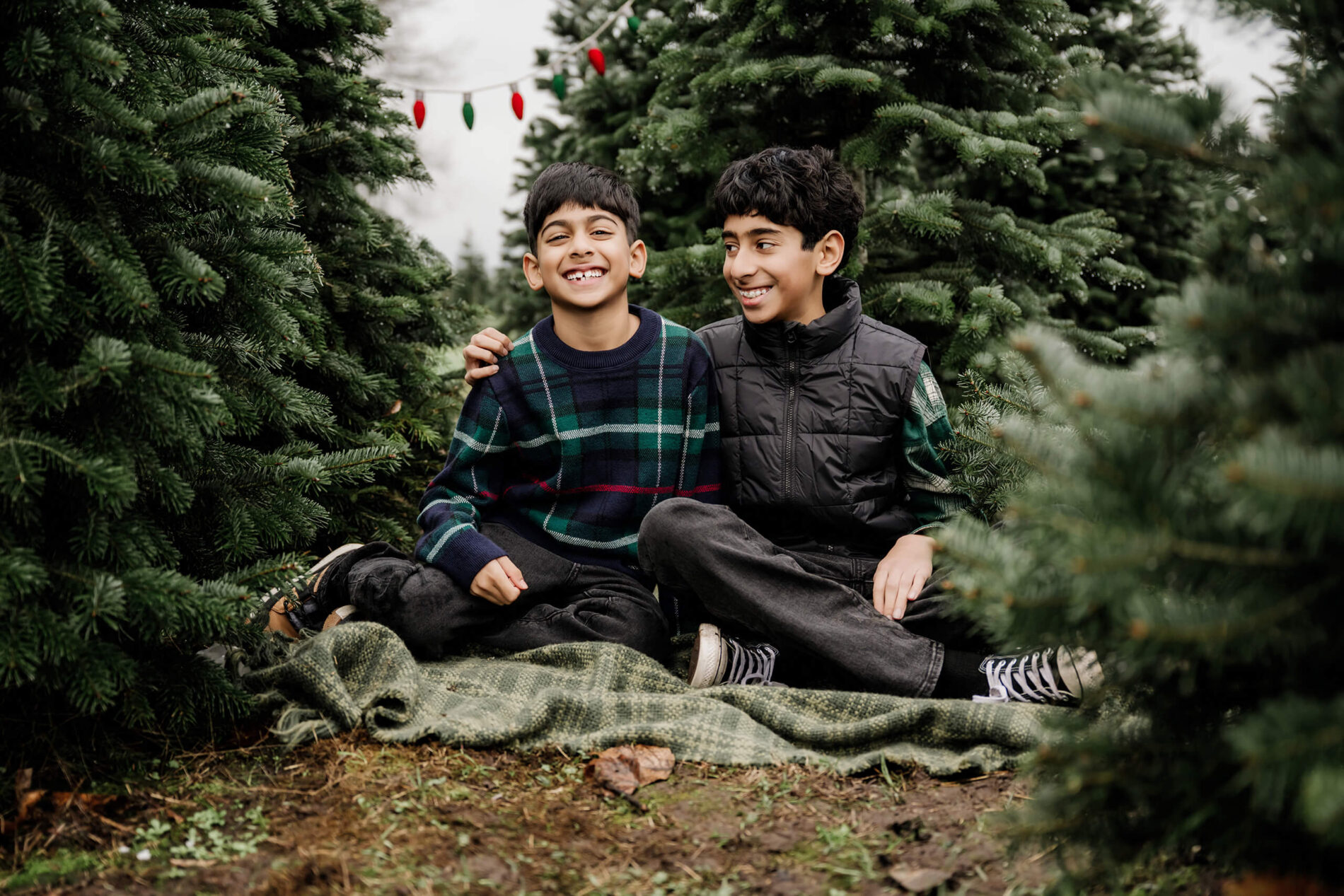 Two brothers sit together on a blanket between evergreen trees, one smiling toward the camera as the other looks at him.