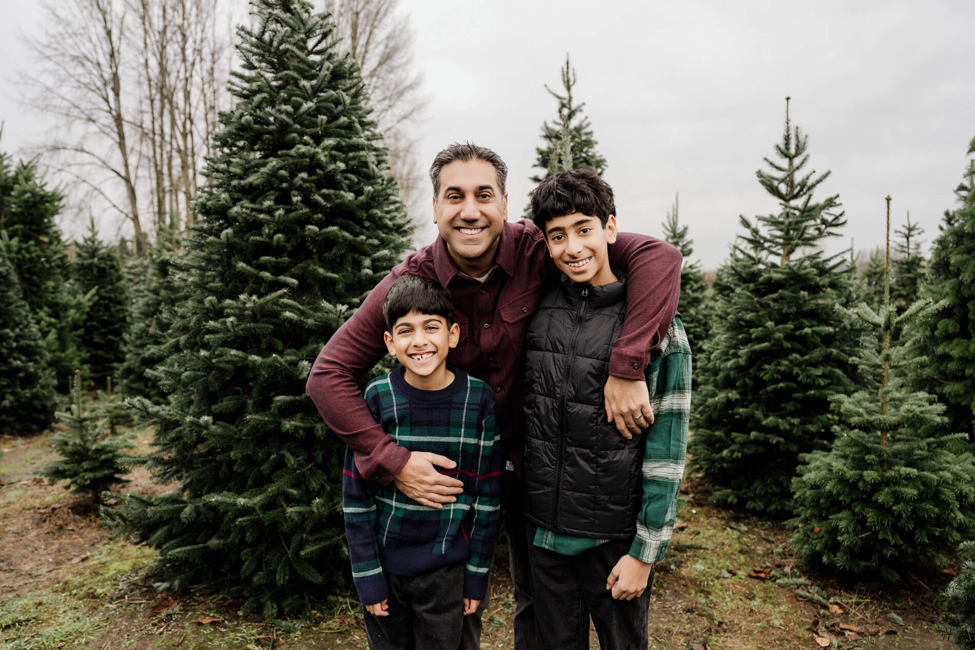 A dad poses with his two sons, an arm around each, among evergreen trees.