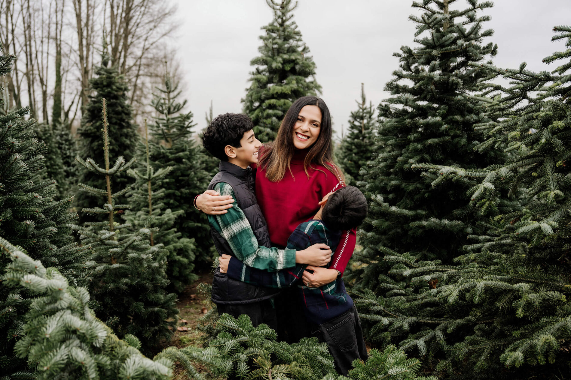 A mom laughs as her two sons hug her between rows of evergreen trees.