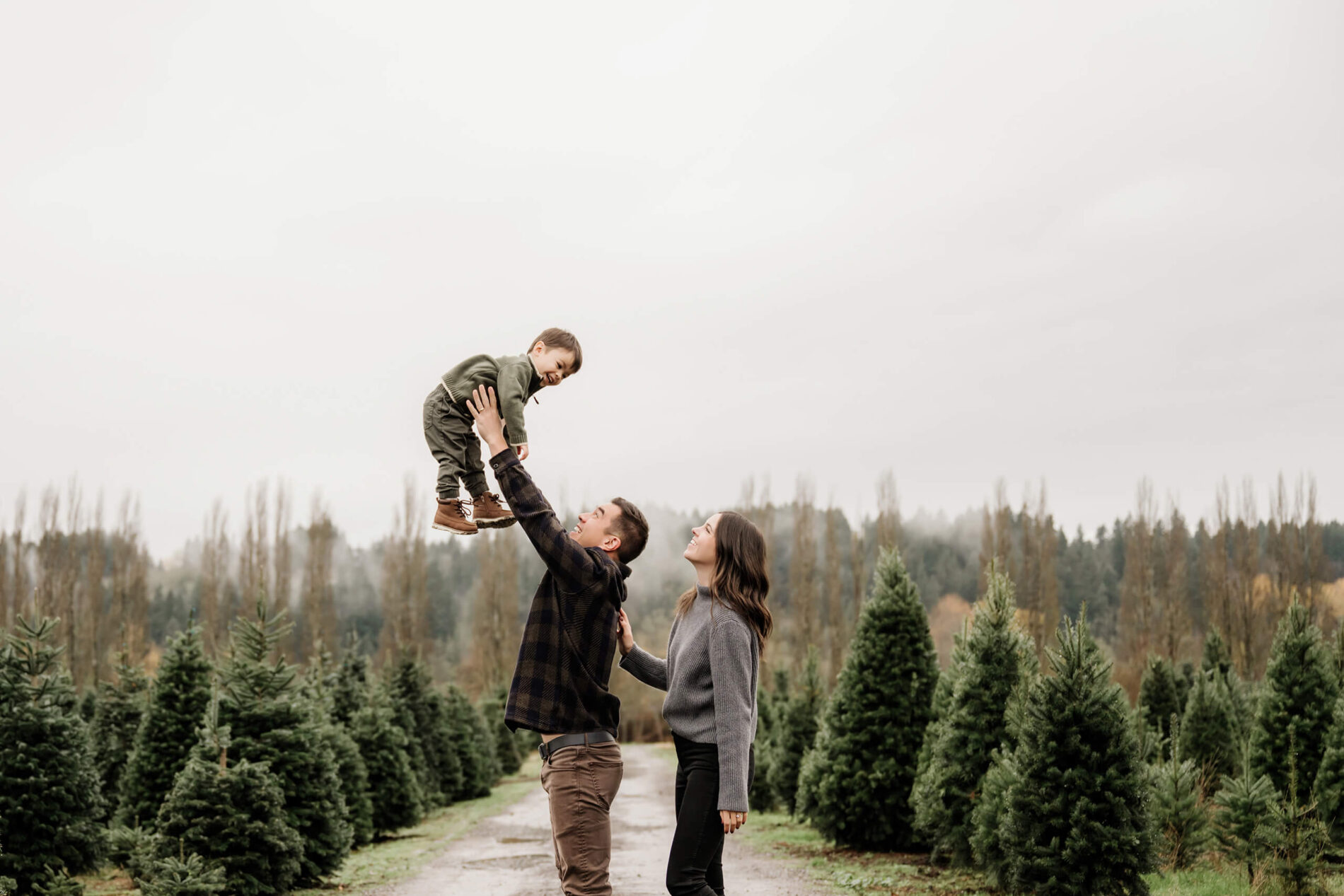 Dad lifts his son high overhead while mom watches along a lane lined with rows of evergreen trees.