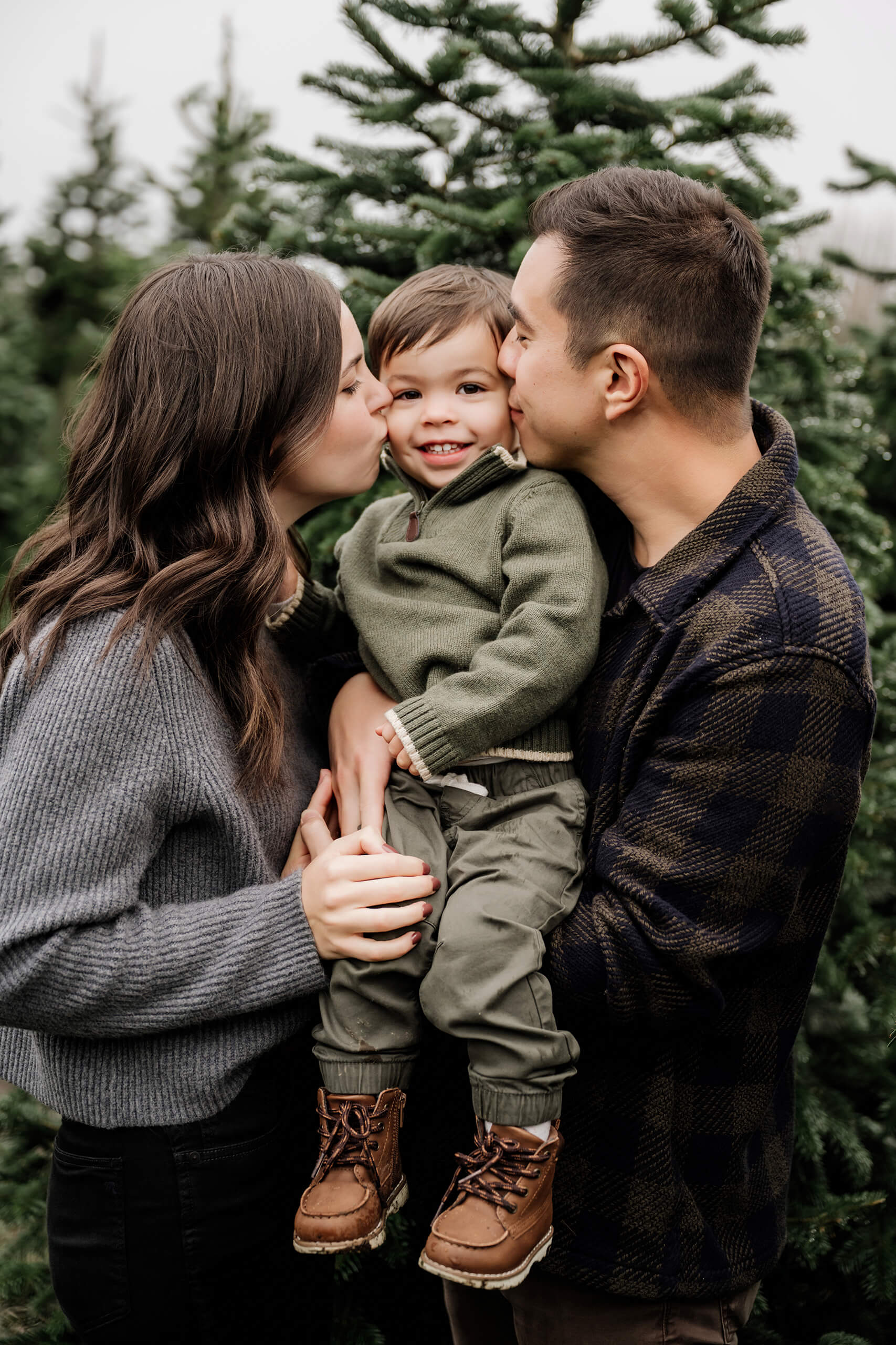 Mom and dad kiss their smiling son on both cheeks in a close portrait among the evergreens.
