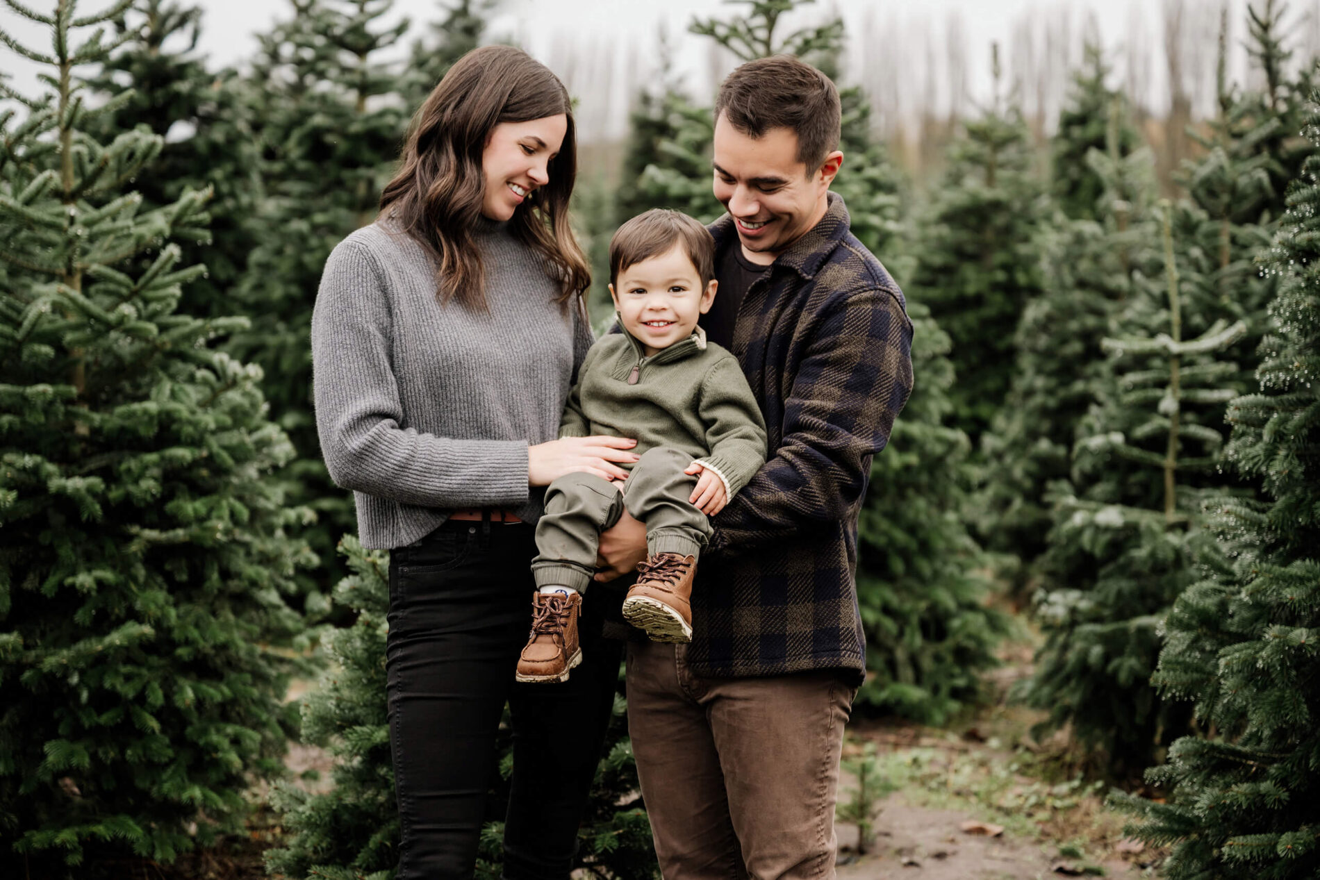 Mom and dad hold their son between them, smiling down at him as he looks at the camera among evergreen trees.