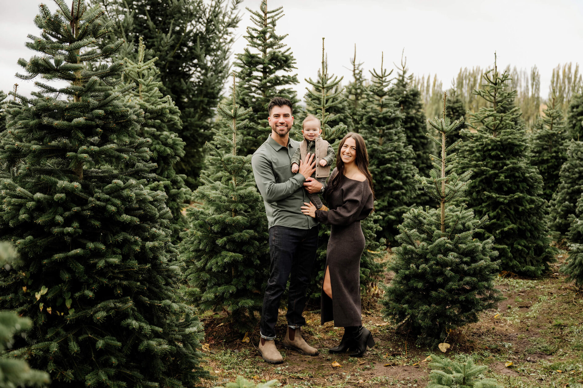 Mom and dad pose with their young son, held in dad’s arms, among rows of evergreen trees.