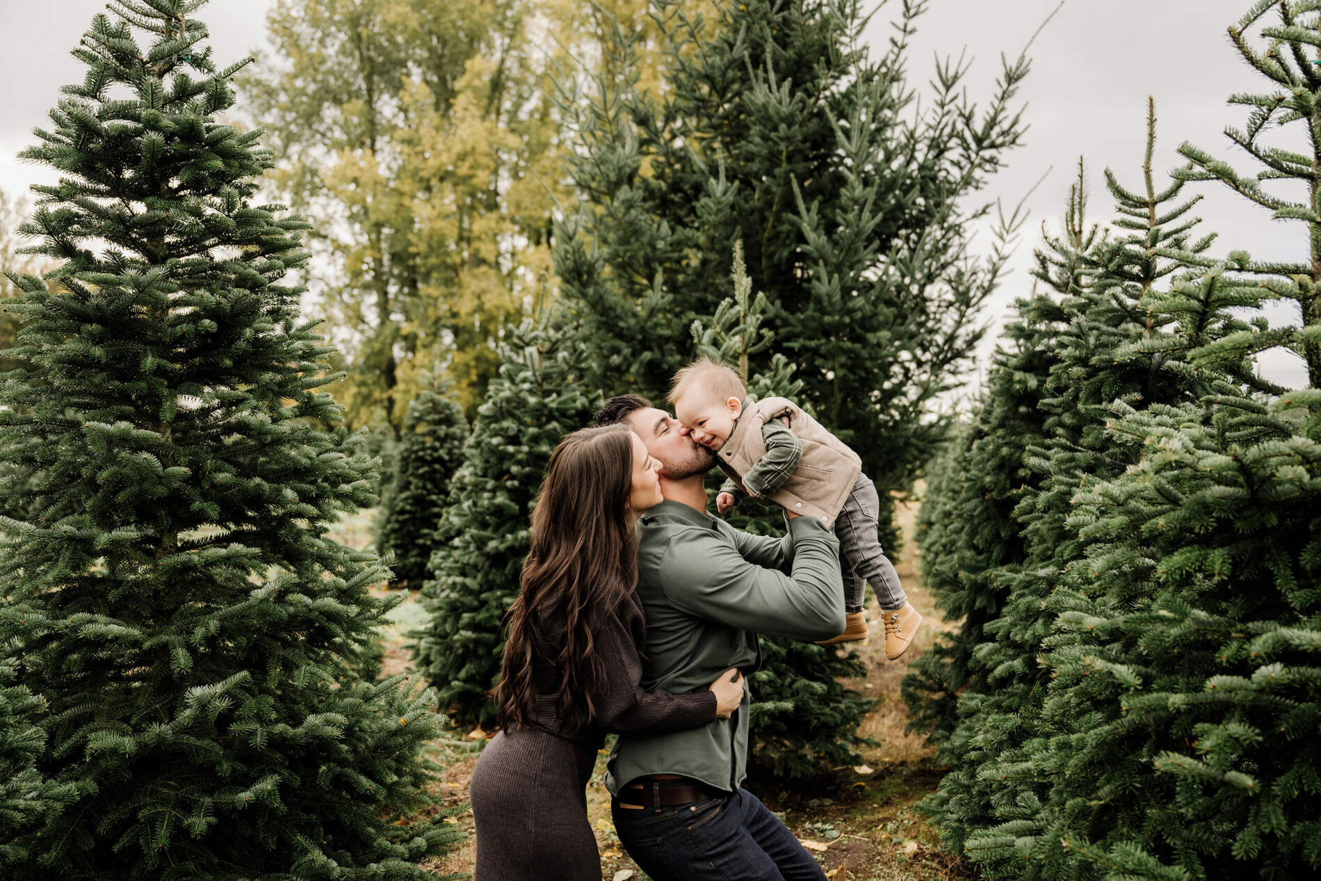 Dad lifts his son playfully as mom stands close beside them between rows of evergreen trees.
