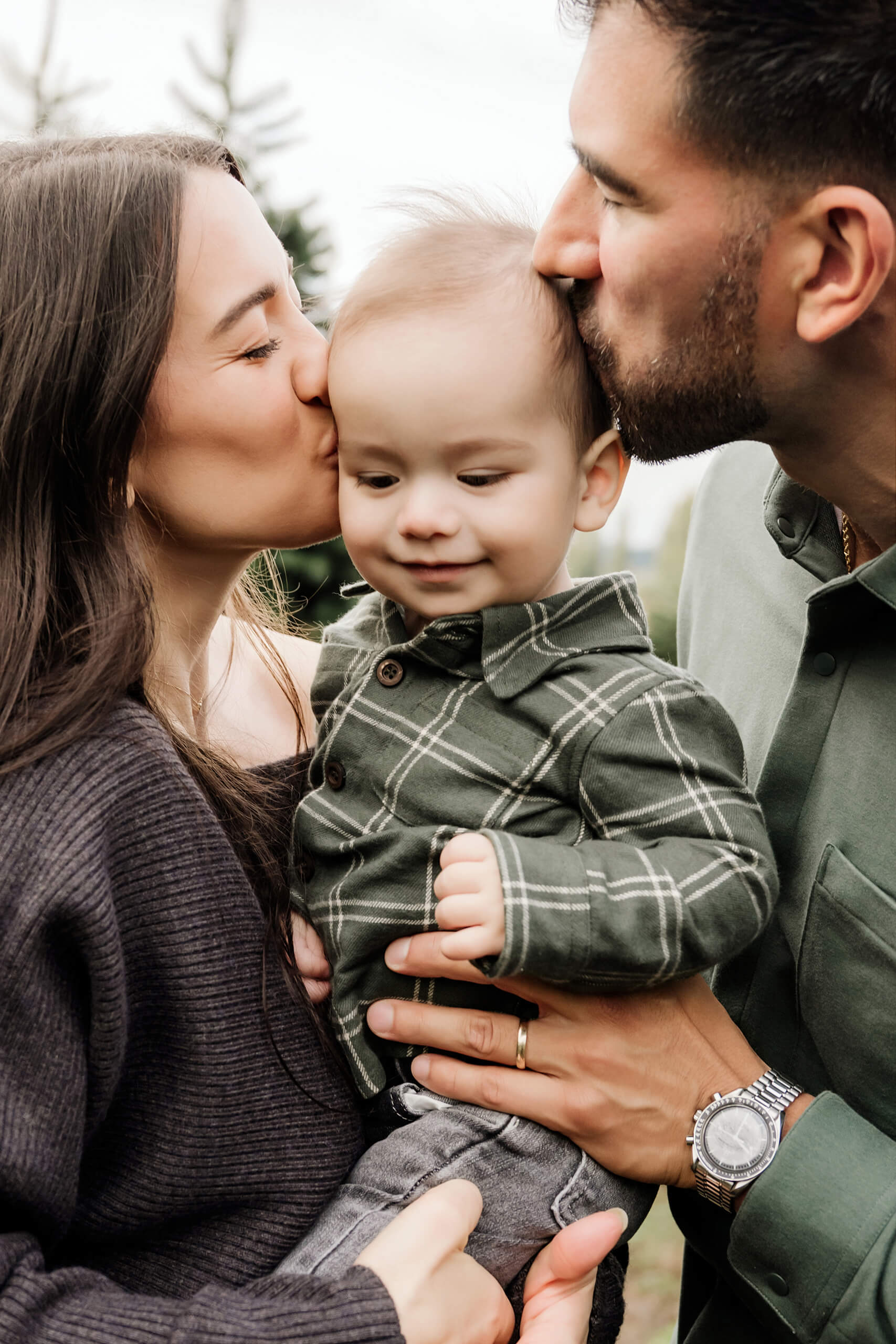 Mom and dad kiss their son from both sides as he smiles between them.