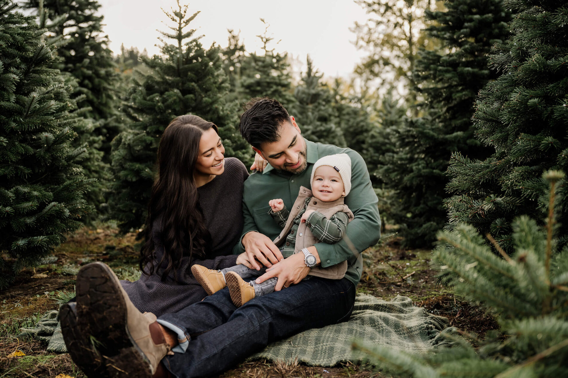 Mom and dad sit together on a blanket as dad holds their smiling son between the evergreen trees.