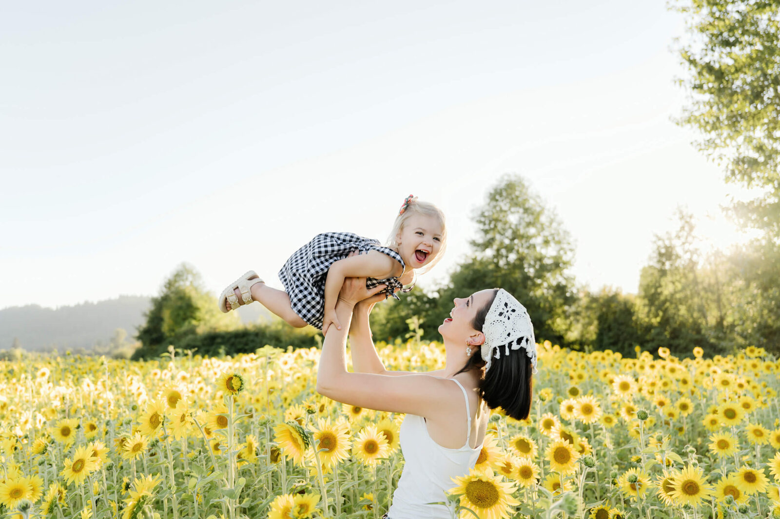 A mother lifting her laughing daughter above a field of yellow sunflowers while looking up at her.