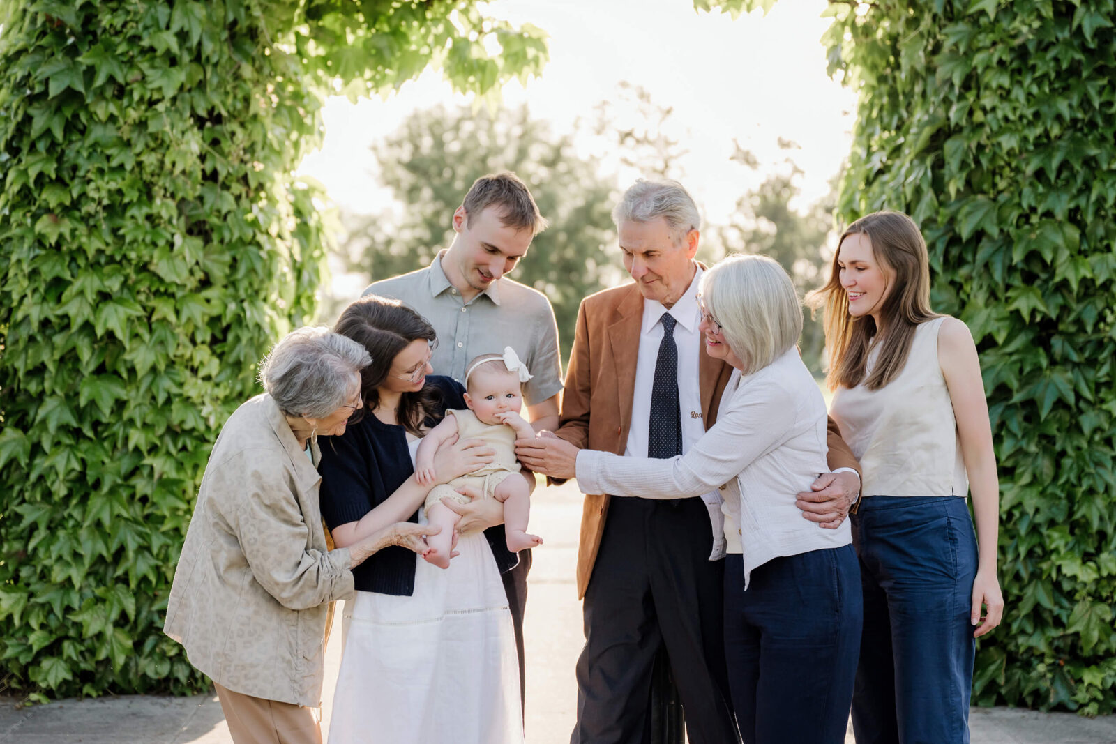A multigenerational family gathered beneath an ivy-covered arch, smiling and leaning toward a baby held in the center.