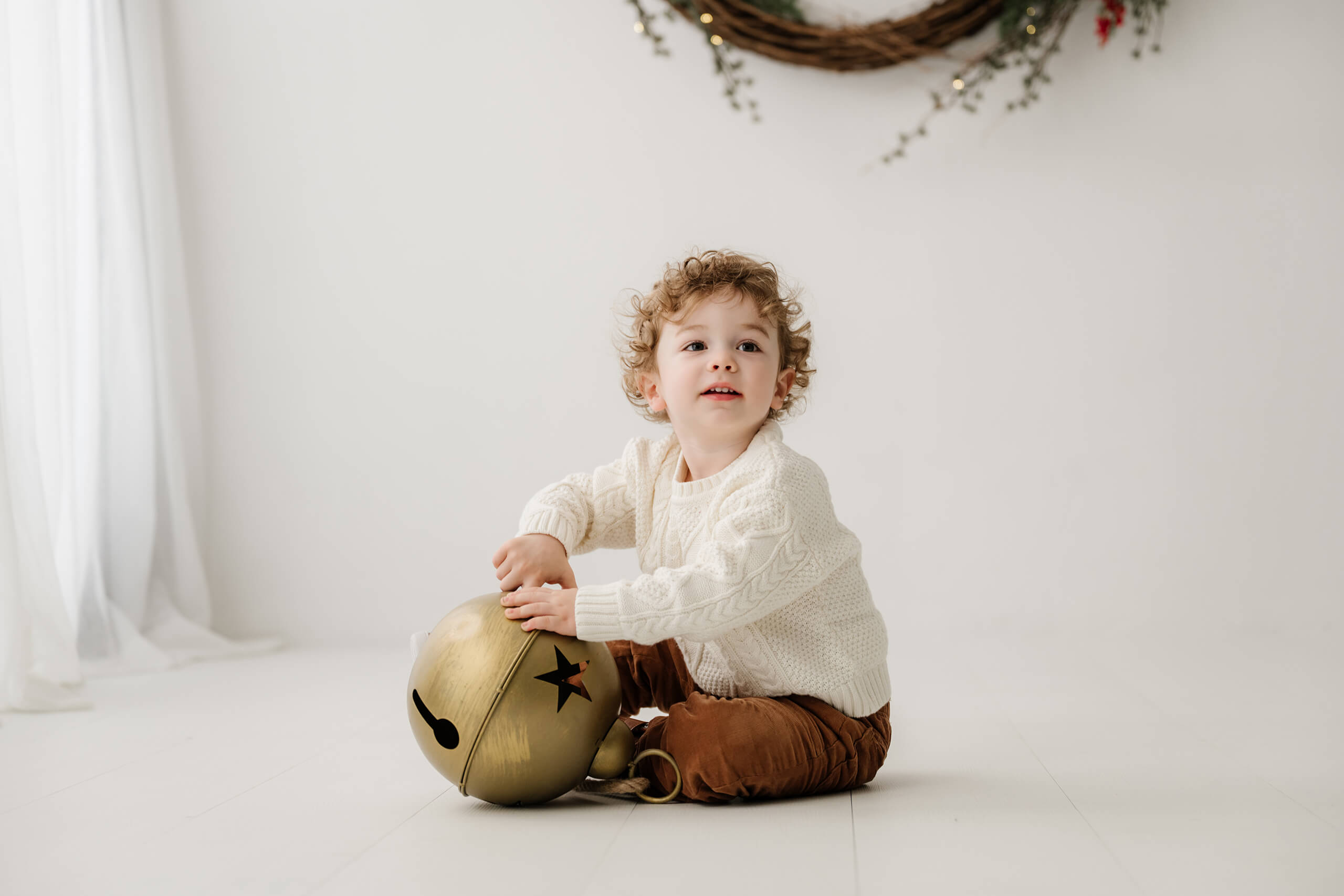 Young boy sitting on the white studio floor with an oversized gold bell beneath the holiday wreath.