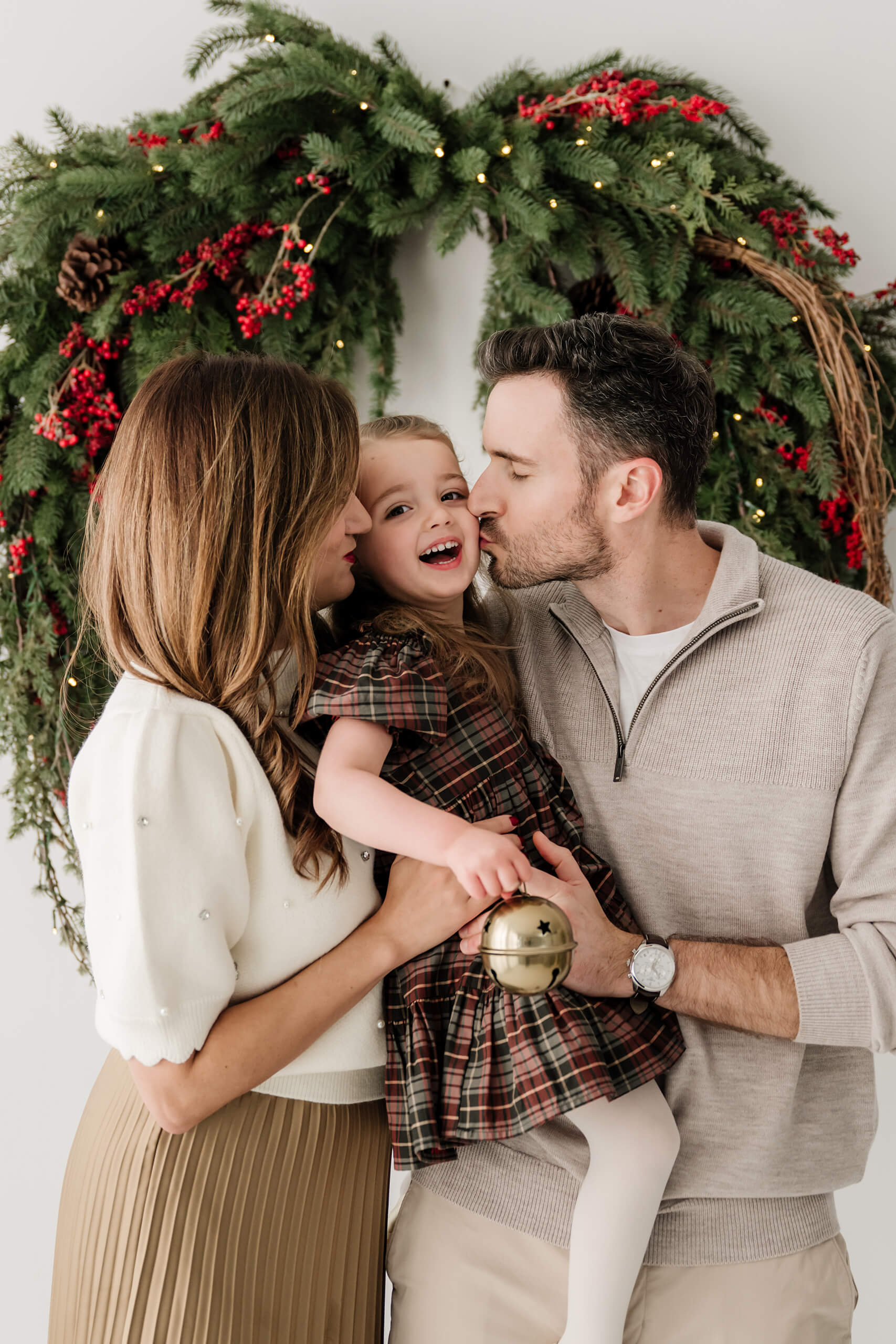 Mom and dad kissing their laughing daughter on the cheeks beneath the evergreen wreath.