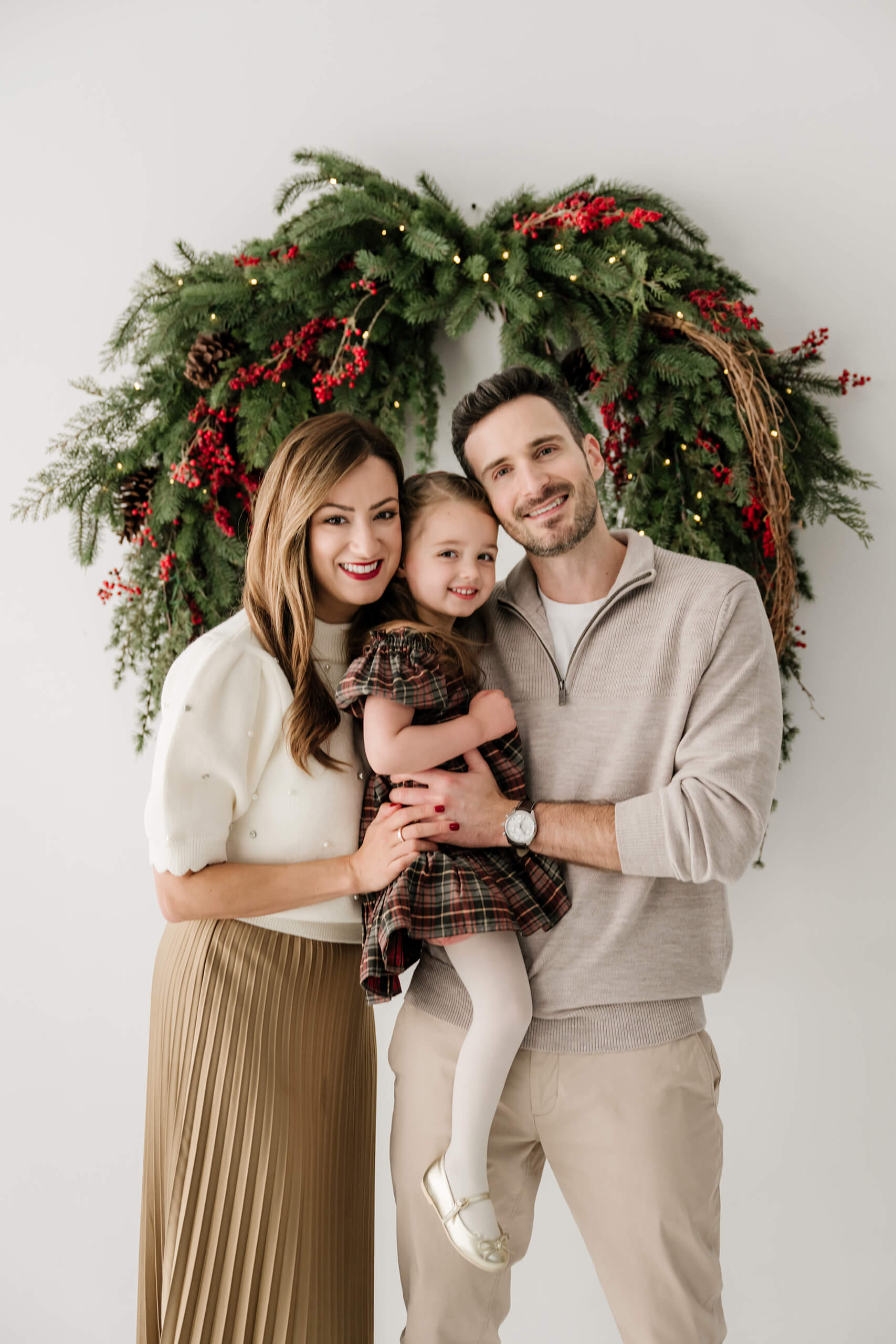 Mom, dad, and their daughter standing close together and looking at the camera beneath the holiday wreath.