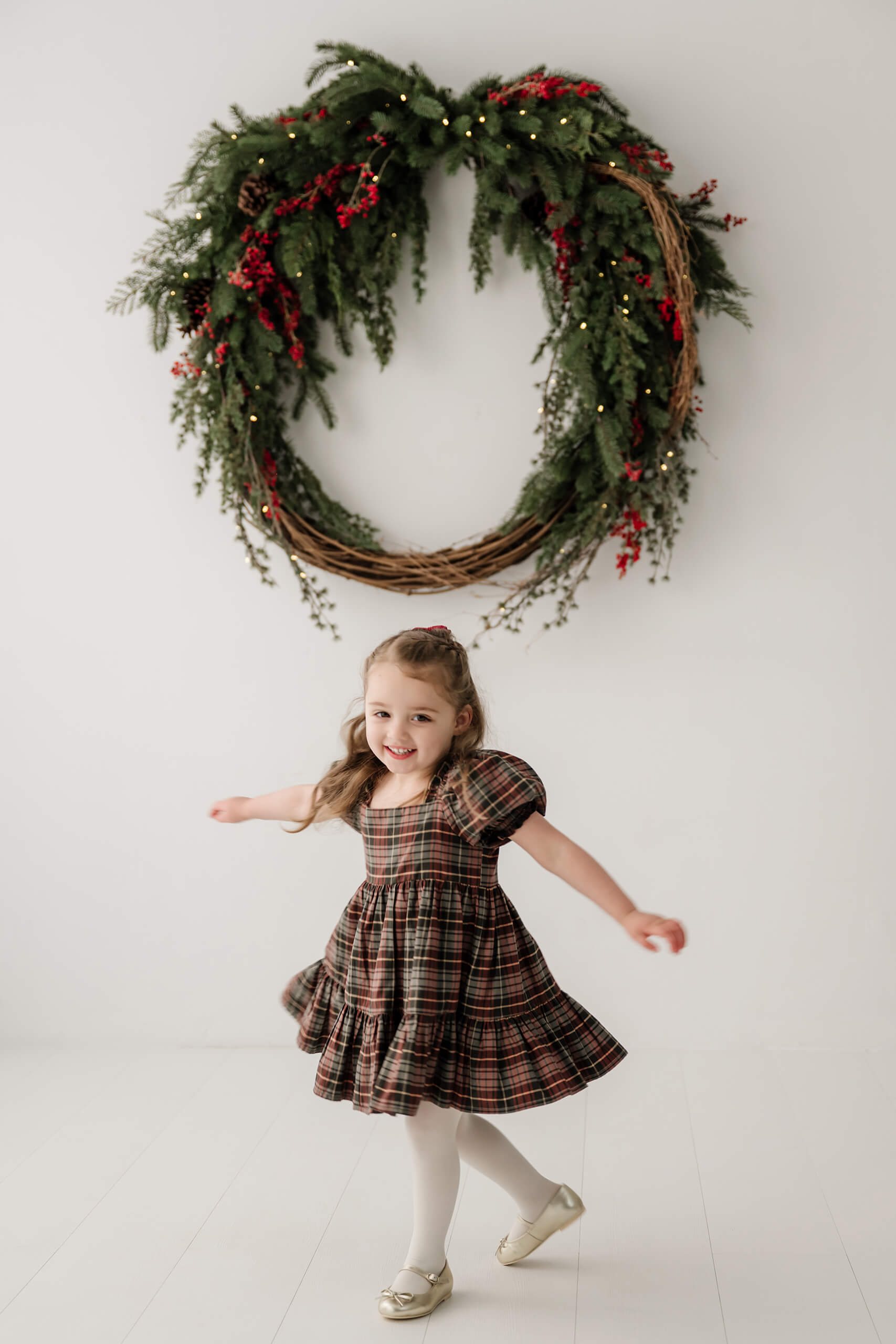 Girl twirling in a plaid dress beneath the oversized evergreen wreath.