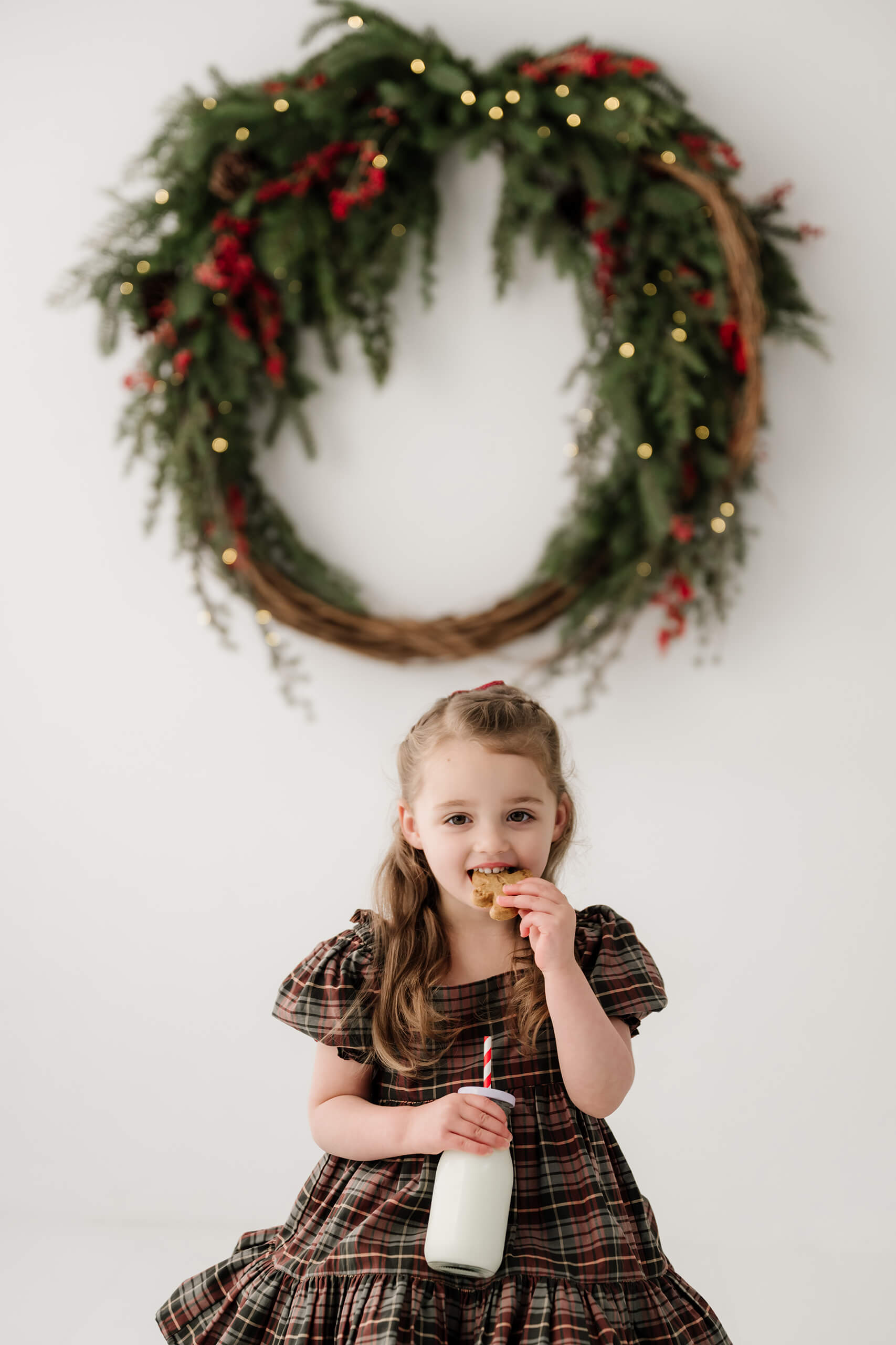 Girl holding a bottle of milk and taking a bite of a cookie, with the holiday wreath softly blurred behind her.