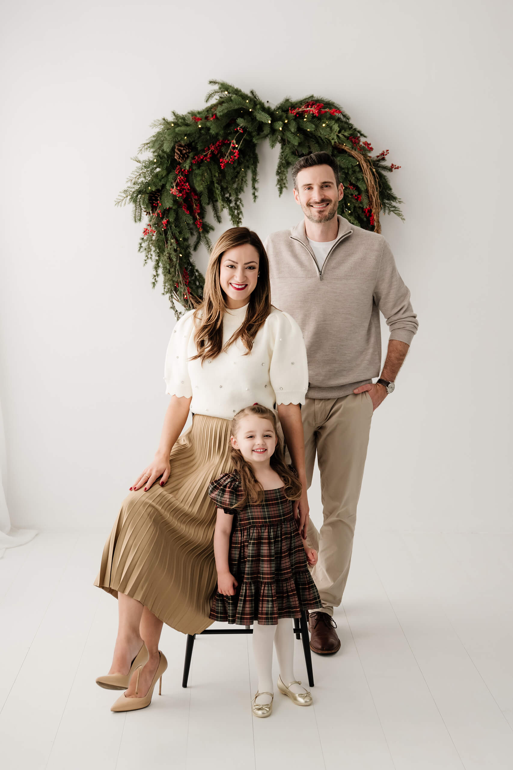 Mom seated with her daughter standing beside her and dad behind them, all looking at the camera beneath the evergreen wreath.