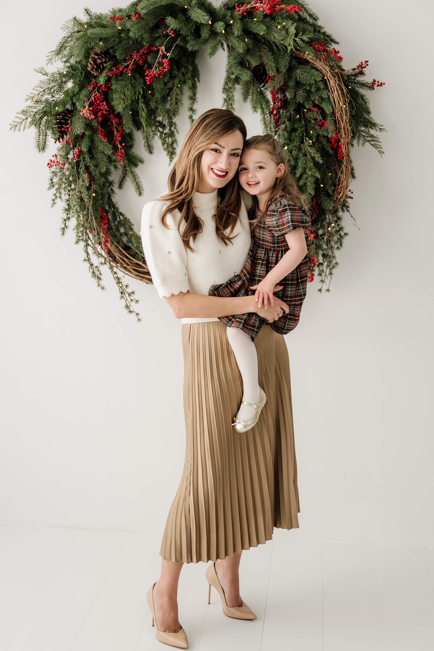 Mom holding her daughter beneath the oversized evergreen wreath, both smiling at the camera.