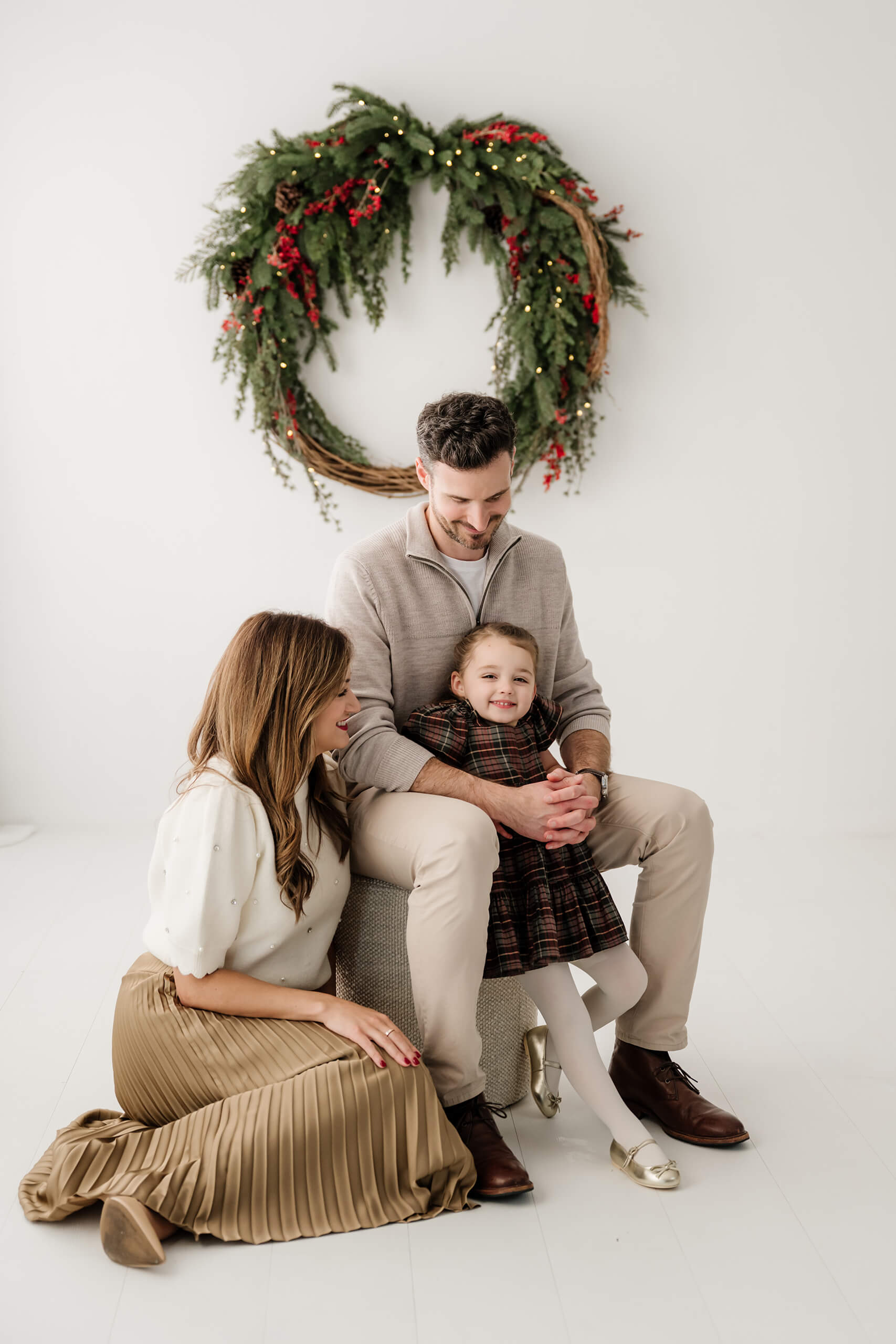 Dad seated with his daughter between his knees while mom sits beside them beneath the evergreen wreath.