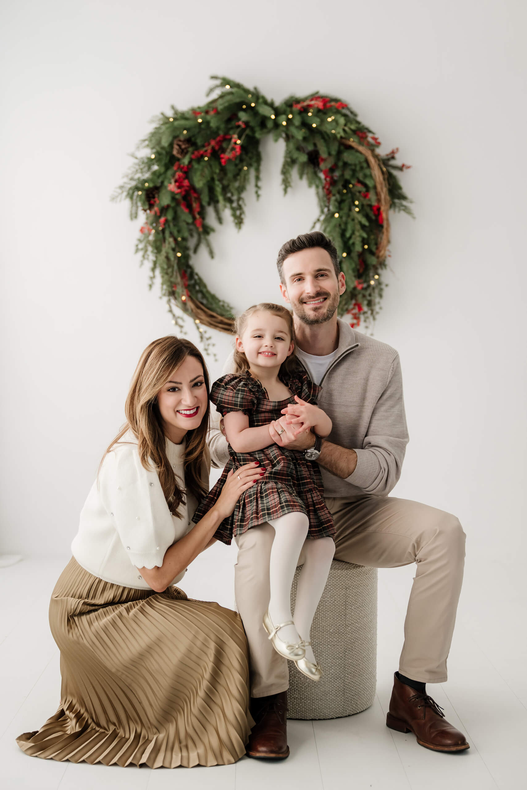 Mom and dad seated with their daughter between them, all looking at the camera beneath the evergreen wreath.
