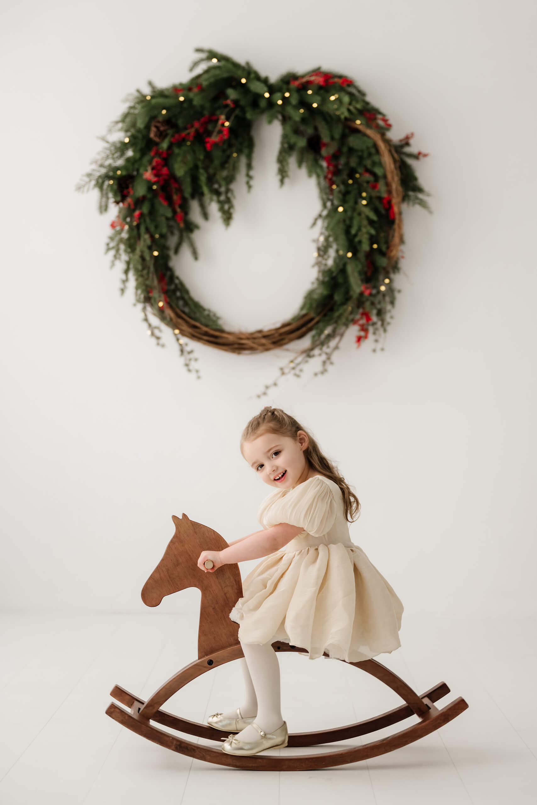 Girl riding a wooden rocking horse beneath the softly blurred evergreen wreath.