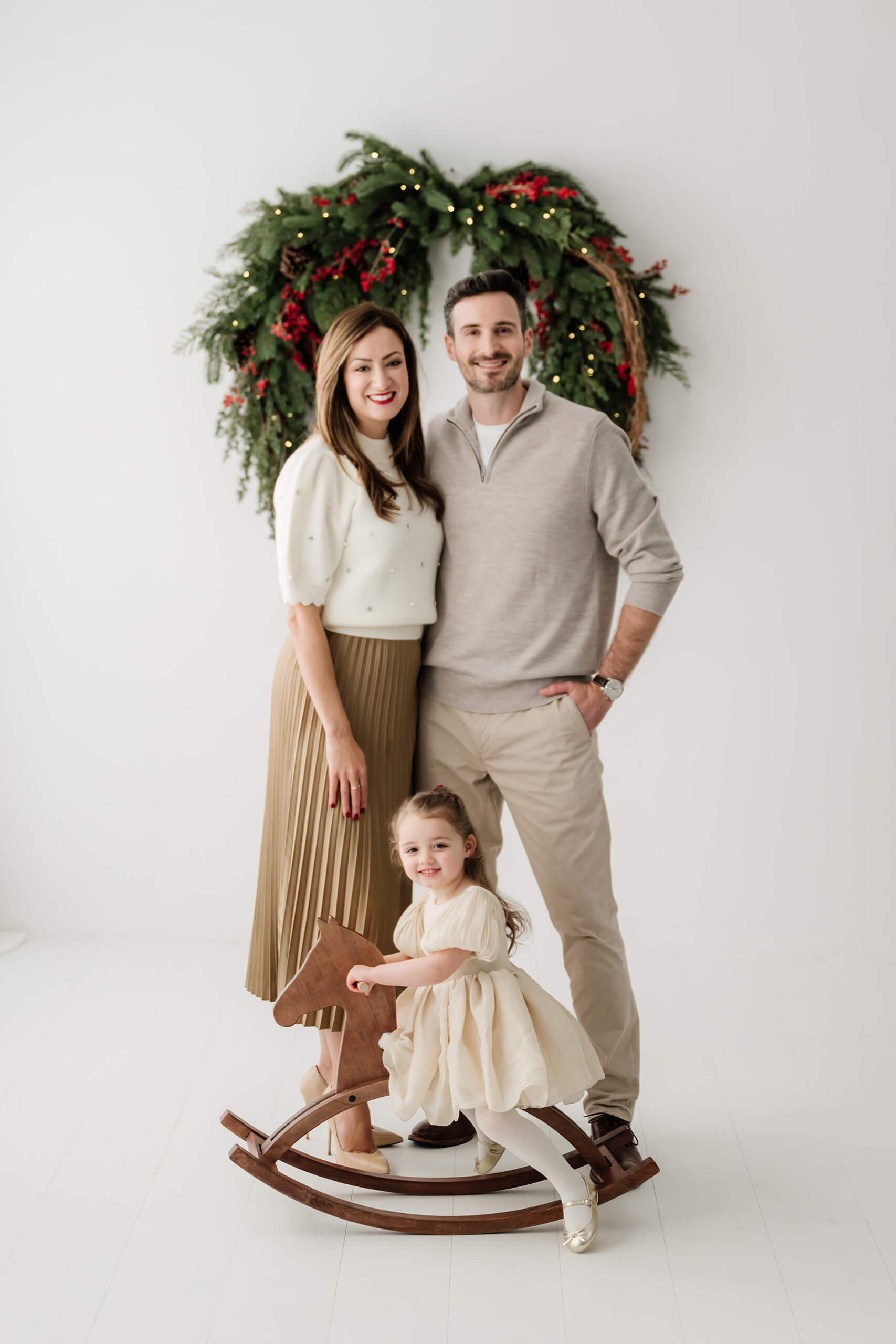 Mom and dad standing behind their daughter as she rides a wooden rocking horse beneath the evergreen wreath.