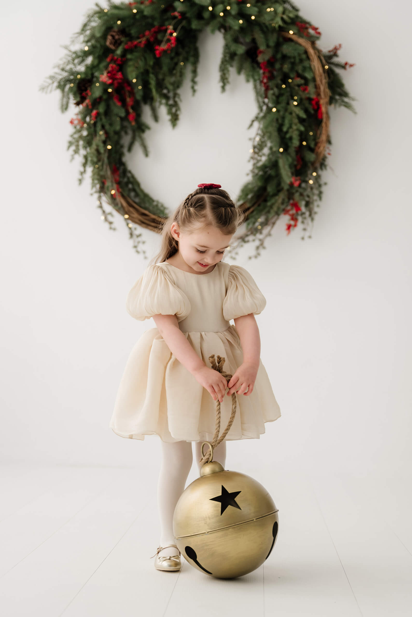 Girl standing and holding the rope of an oversized gold bell beneath the evergreen wreath.