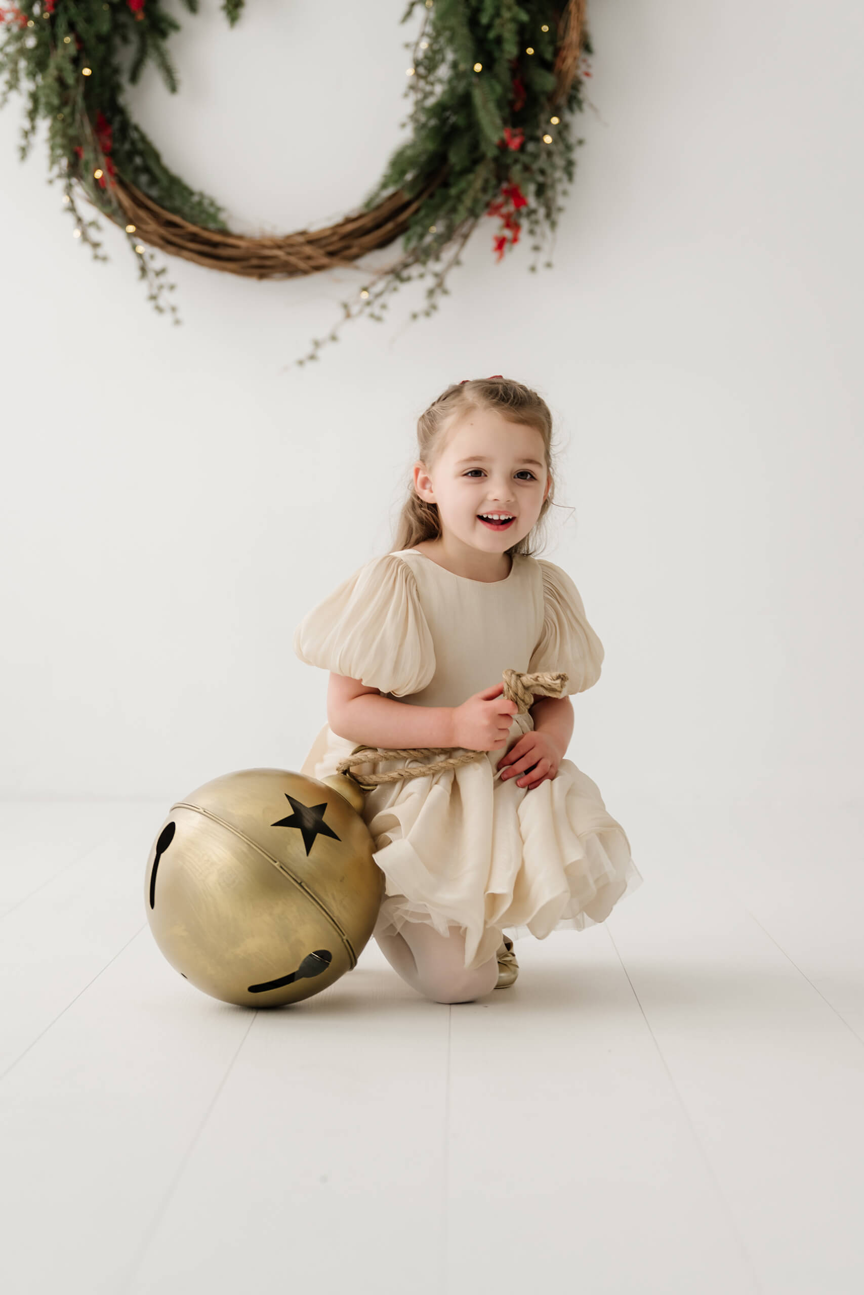 Girl kneeling beside an oversized gold bell beneath the holiday wreath.