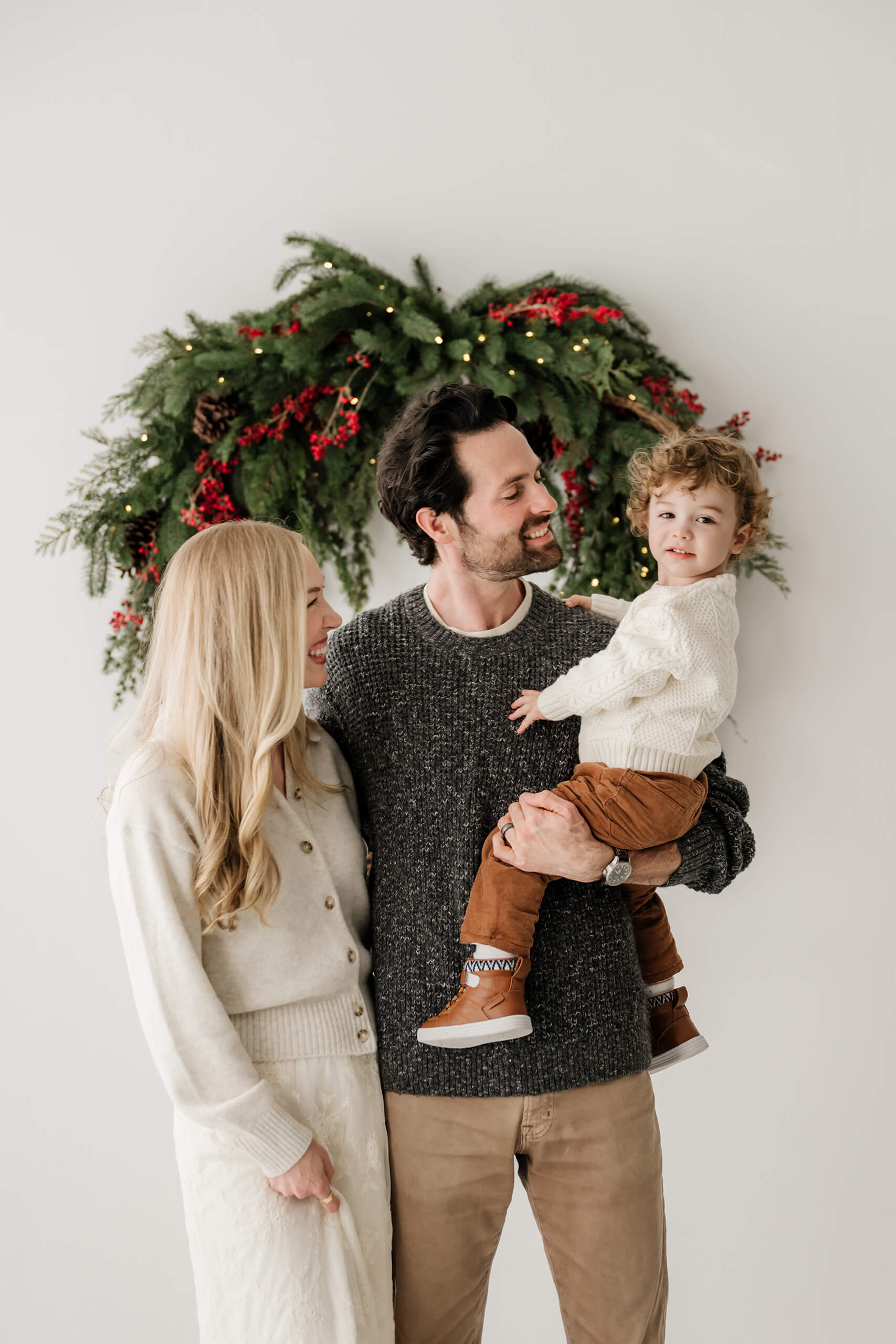 Dad holding his son as mom and dad look toward him beneath the holiday wreath.