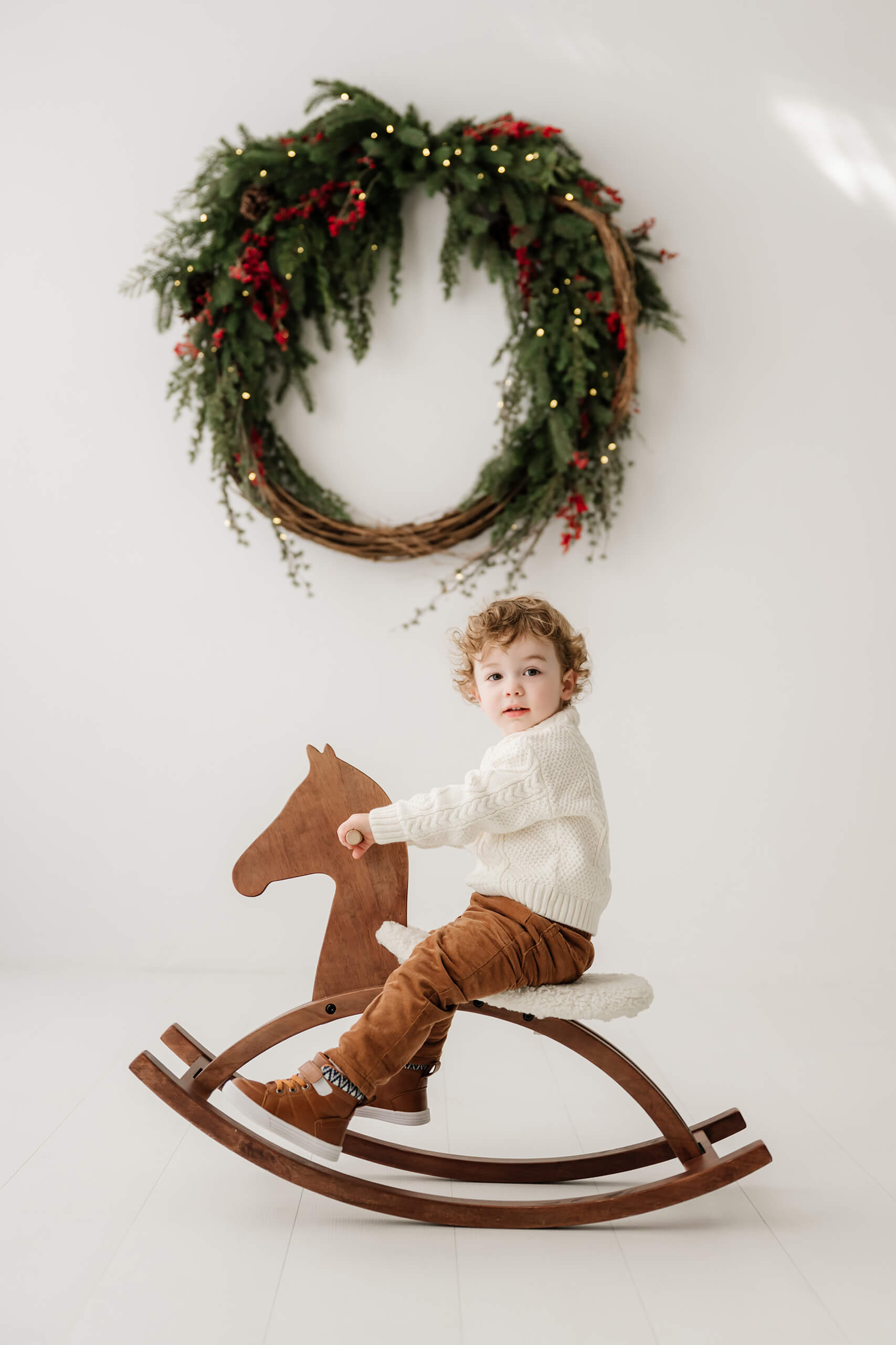 Young boy riding a wooden rocking horse and looking at the camera beneath the evergreen wreath.