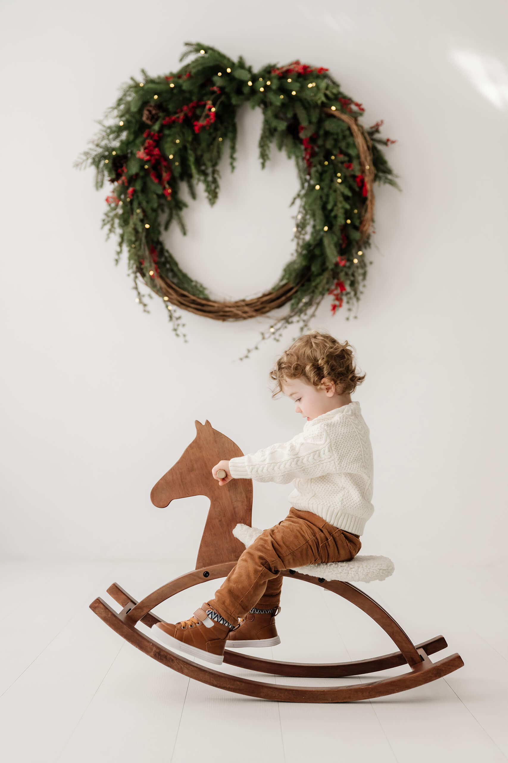 Young boy looking down while riding a wooden rocking horse beneath the holiday wreath.