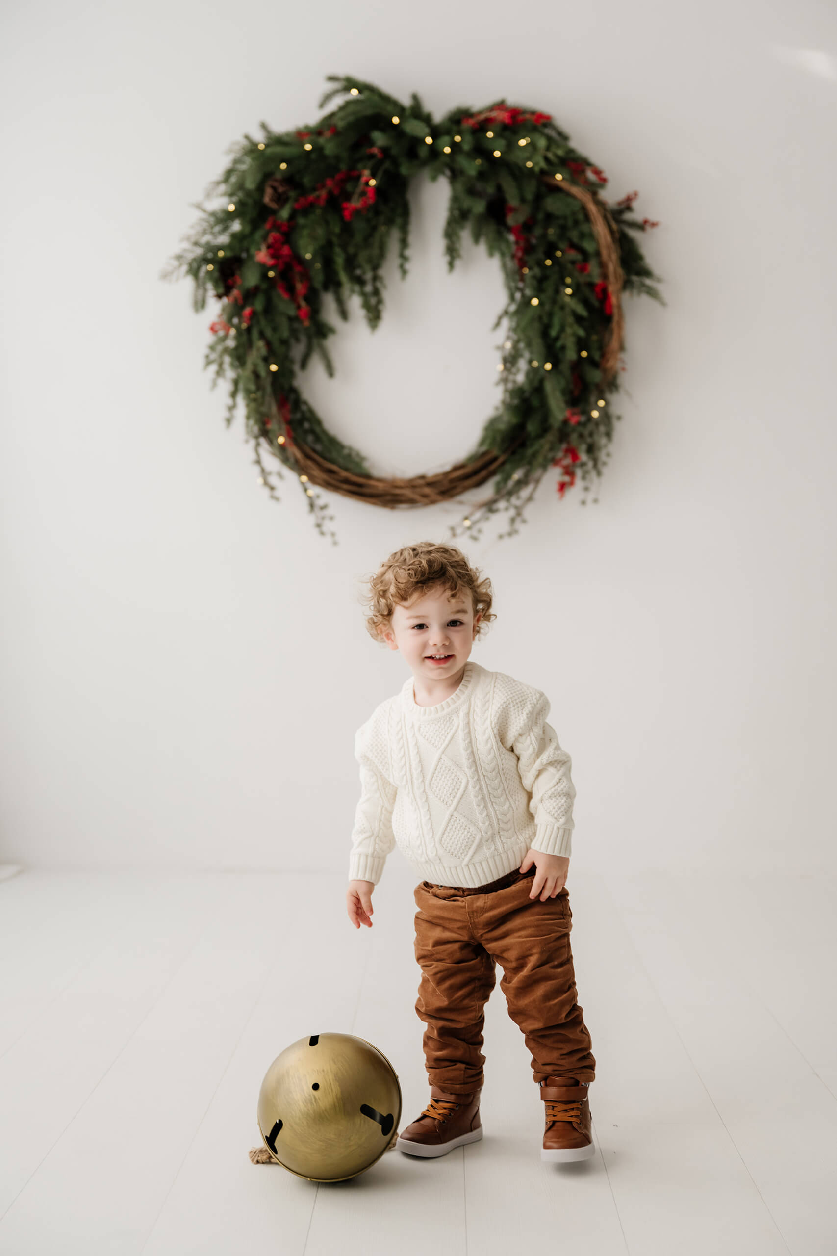 Young boy standing beside an oversized gold bell beneath the evergreen wreath.