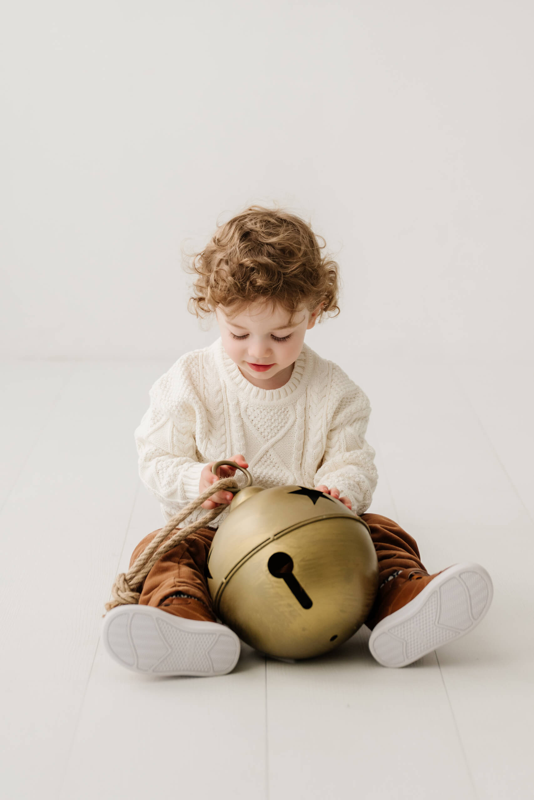 Young boy sitting on the white studio floor and examining an oversized gold bell.