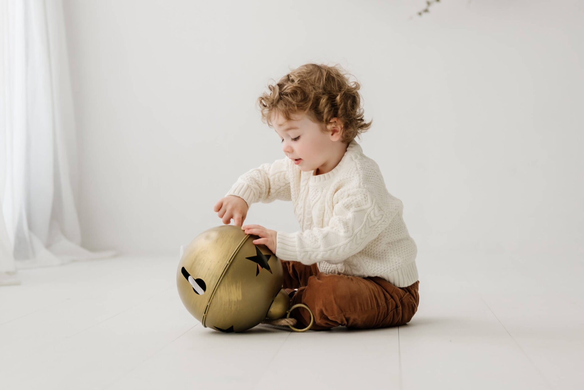 Young boy sitting on the white studio floor and playing with an oversized gold bell.