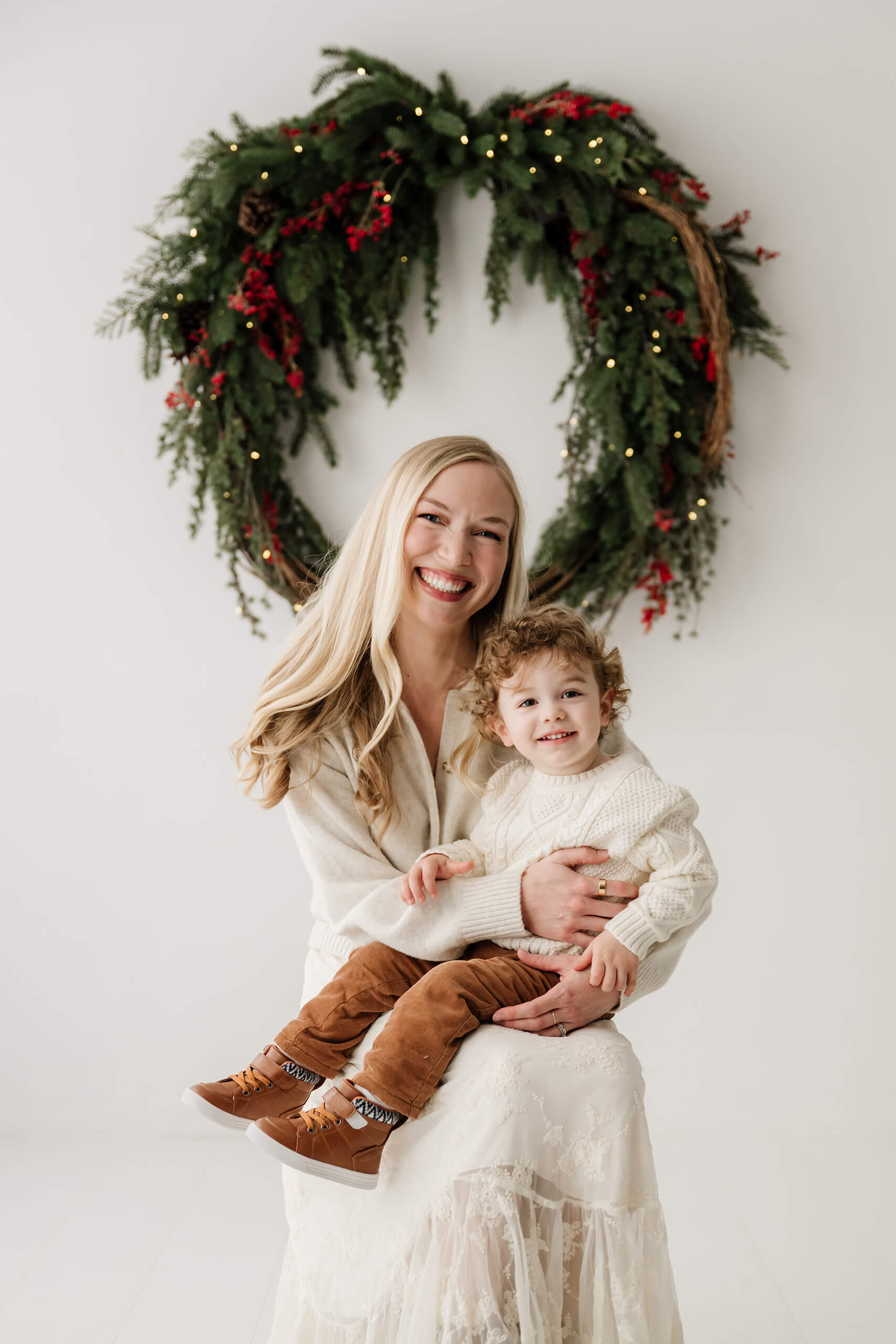 Mom holding her young son as they smile at the camera beneath the evergreen wreath.