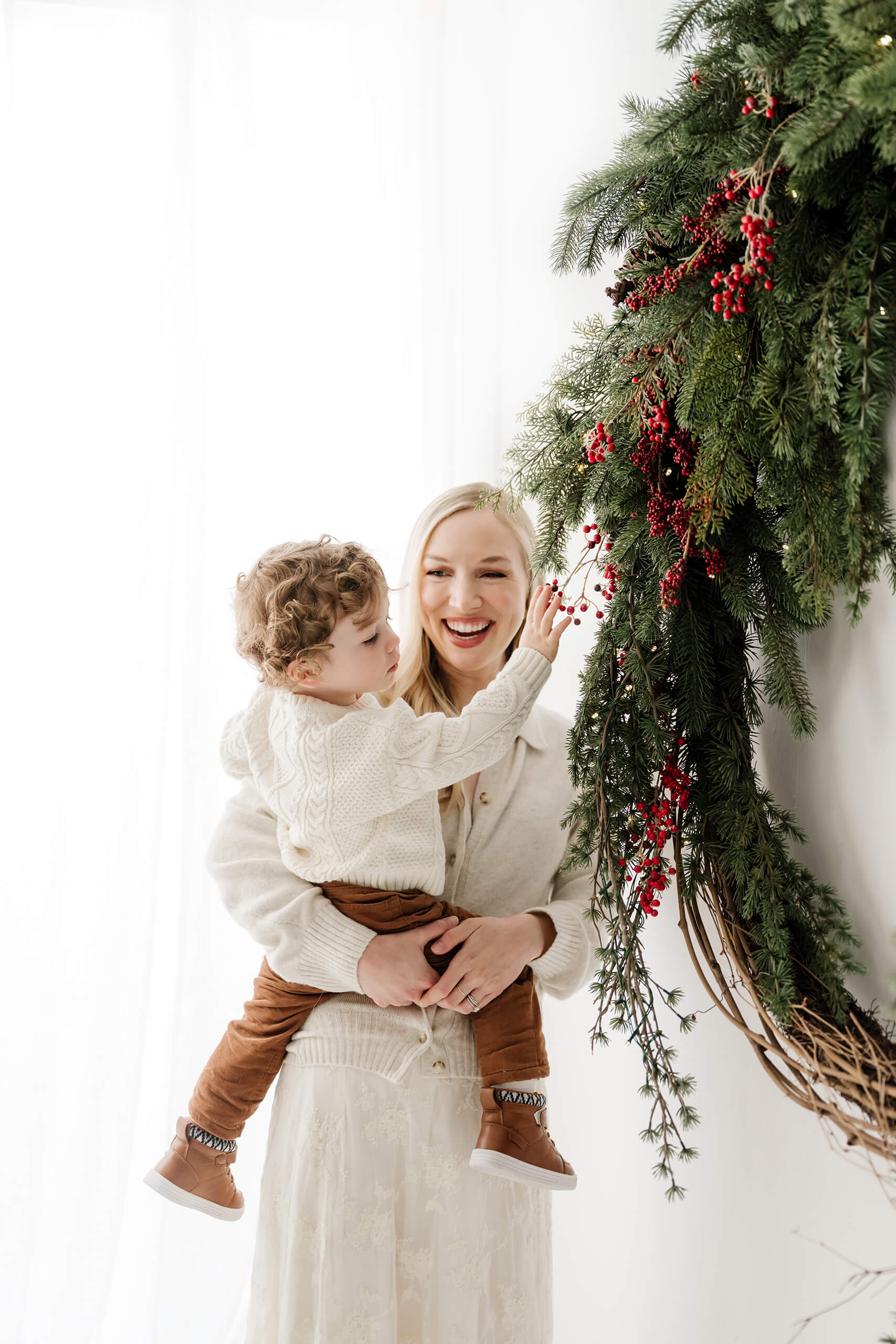 Mom holding her son as he reaches toward the red berries and greenery of the holiday wreath.
