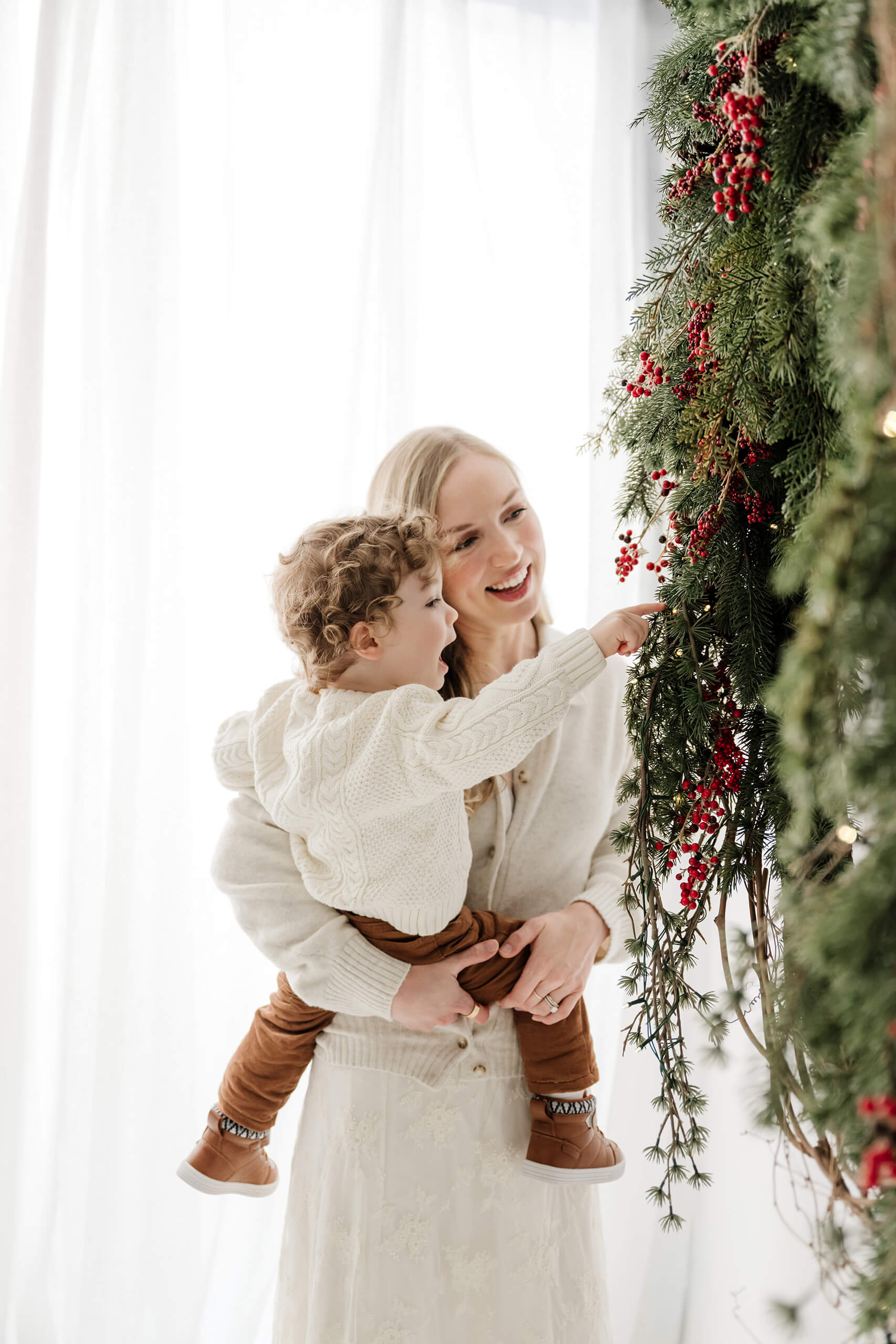 Mom holding her son as he points toward the evergreen wreath and holiday lights.