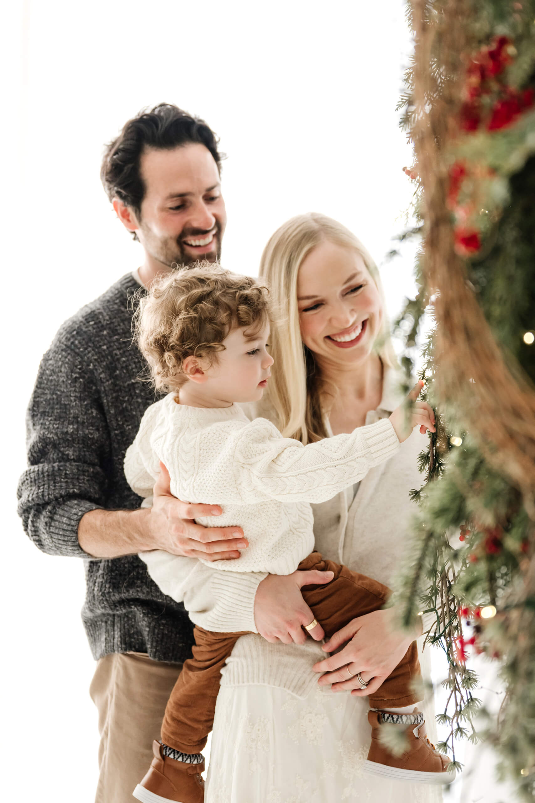 Mom and dad watching their young son explore the greenery and red berries on the holiday wreath.