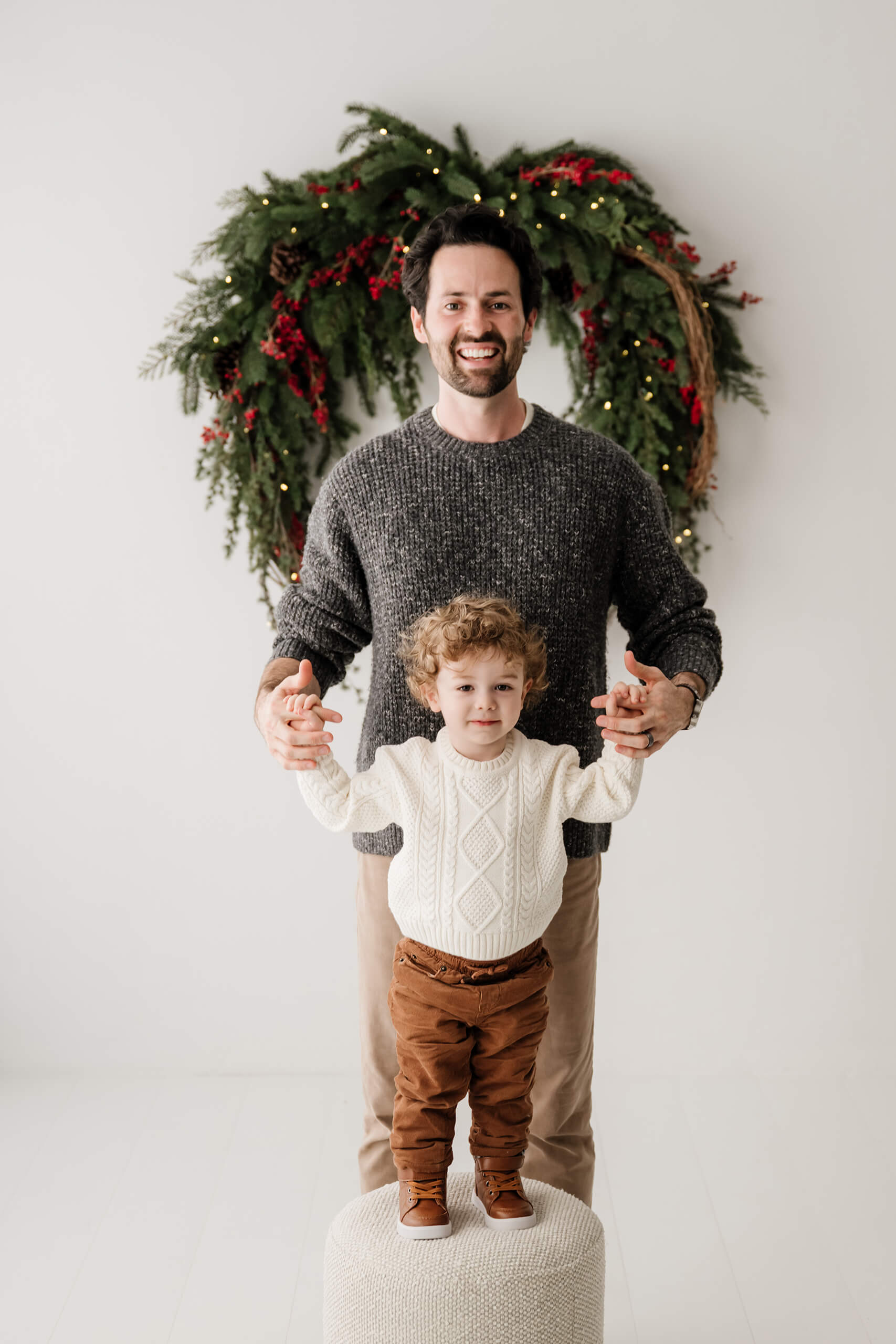 Dad holding his son’s hands as the boy stands on a small ottoman beneath the evergreen wreath.