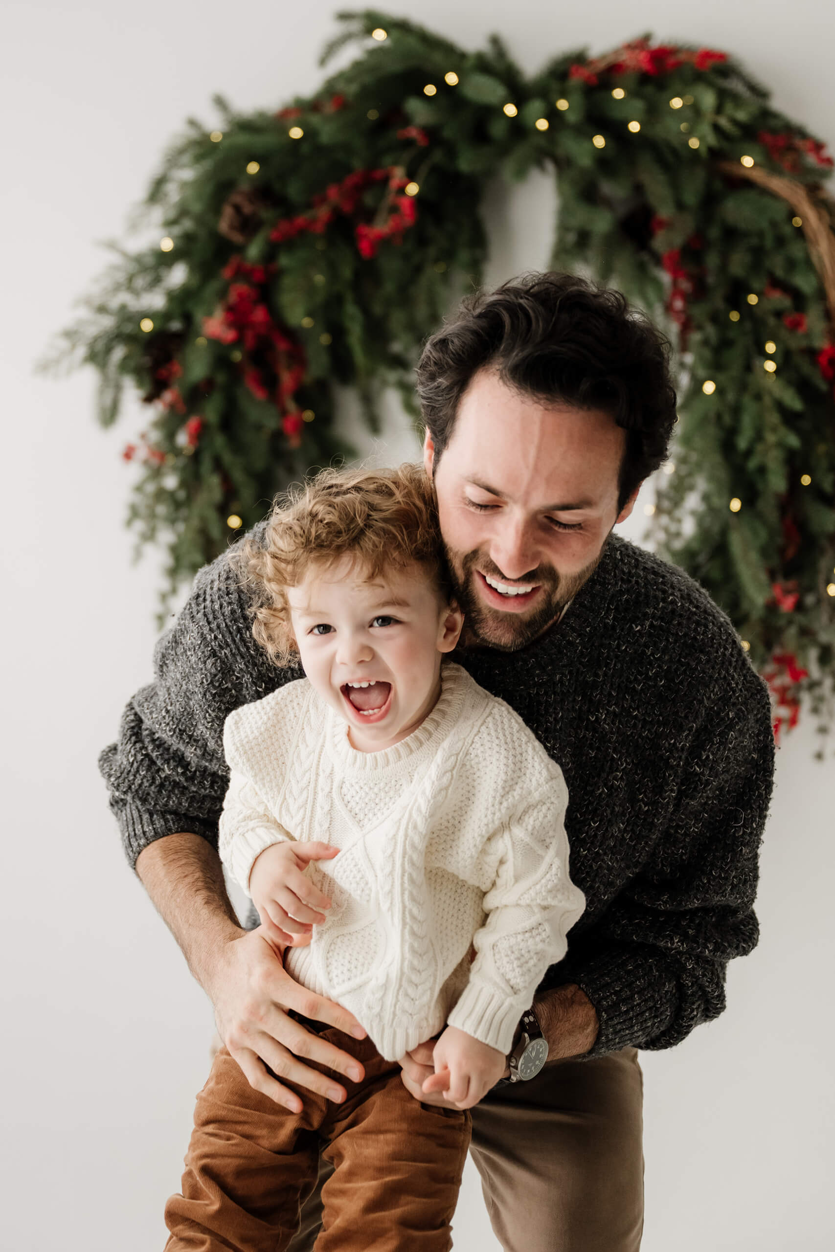 Dad leaning close to his laughing son beneath the softly blurred evergreen wreath.