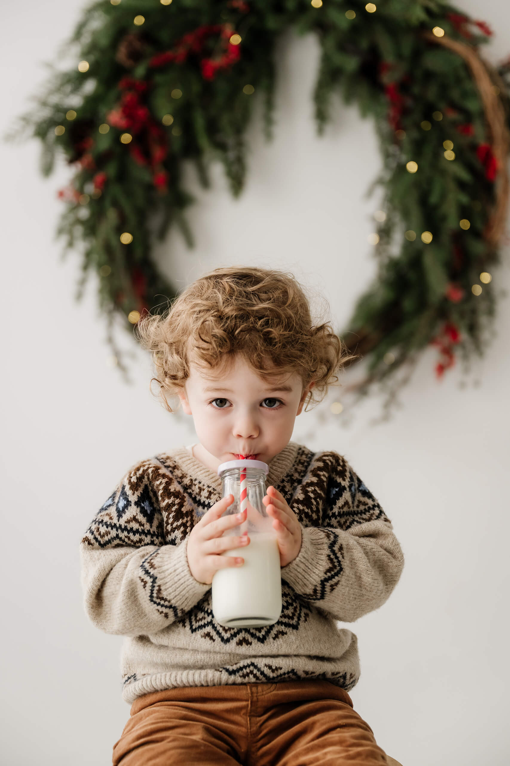 Young boy drinking milk through a striped straw with the holiday wreath softly blurred behind him.